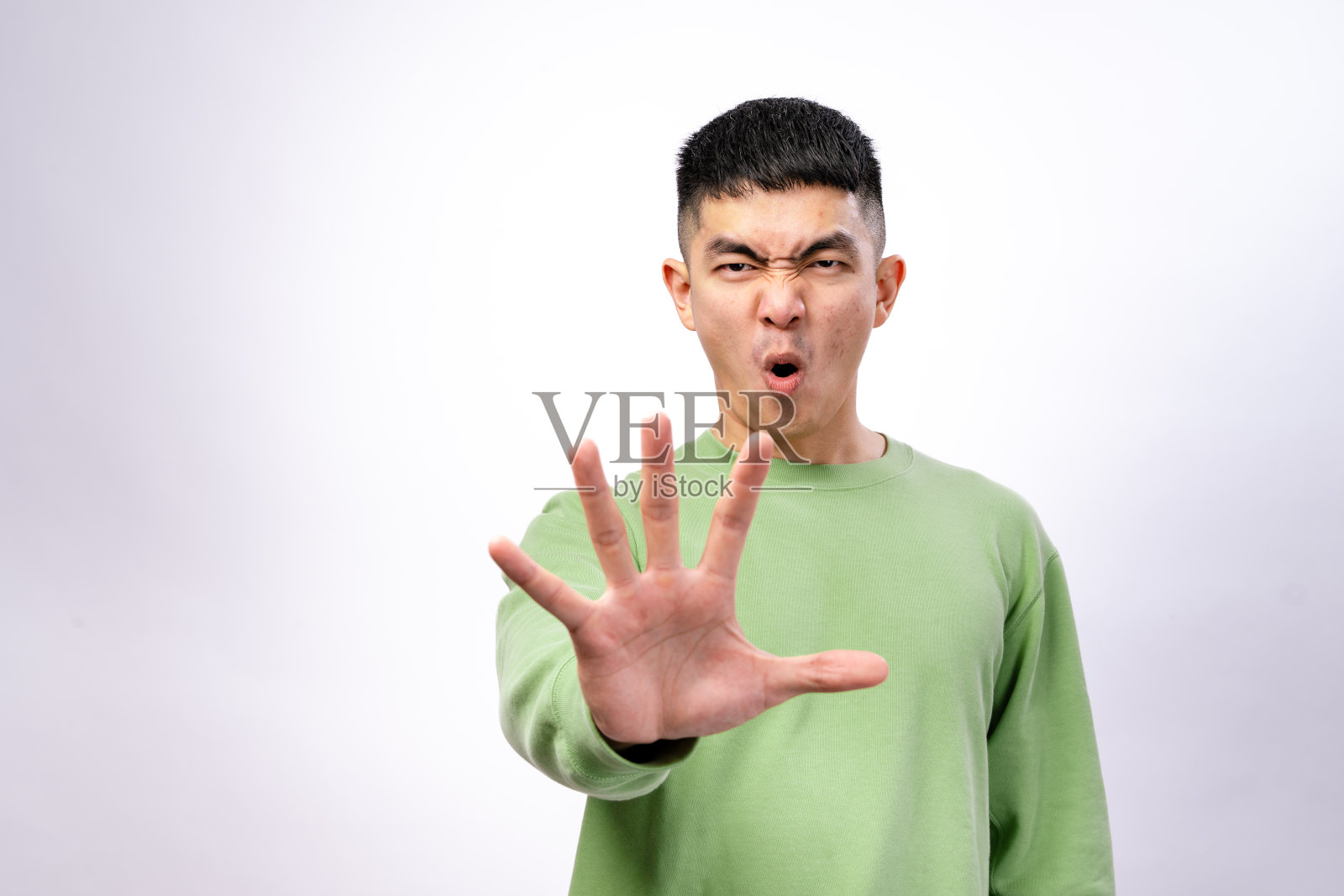 一位穿着绿色卫衣的亚洲男子站在白色背景前,面露严肃表情,伸出手做出停止的手势,表示拒绝、警告或设定界限。照片摄影图片