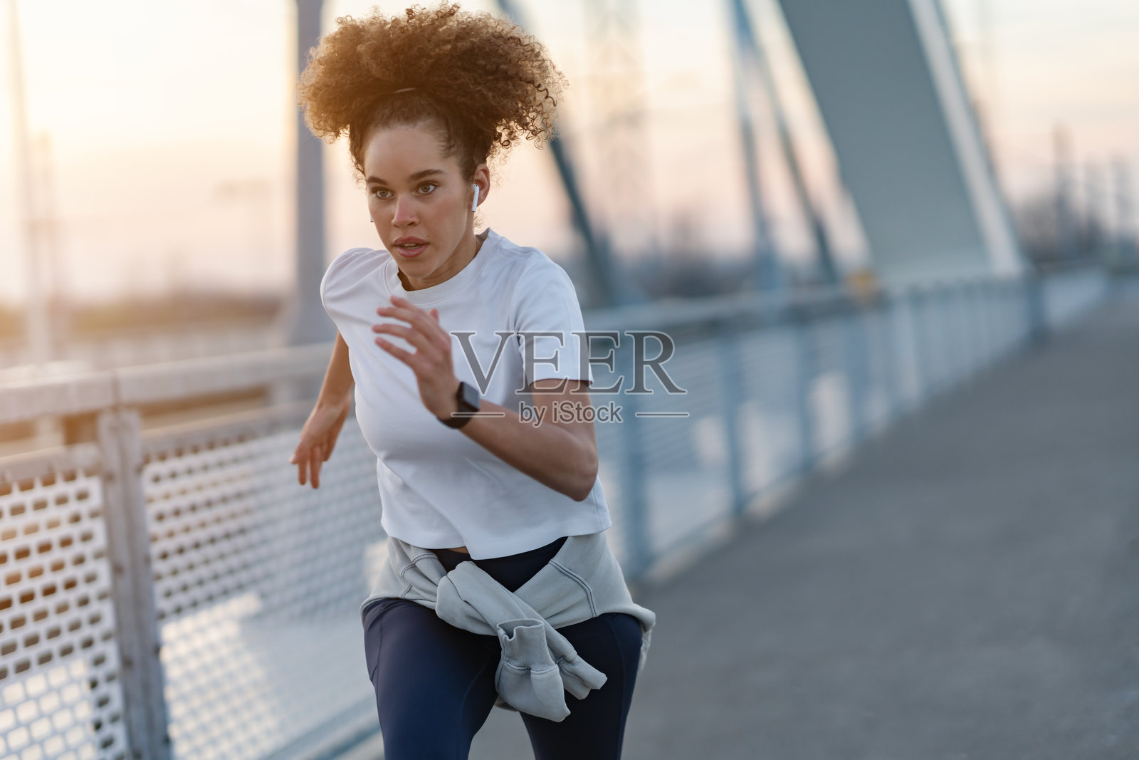 一位决心坚定的年轻女性在现代桥梁上进行户外锻炼,奔跑着。她穿着白色T恤、紧身裤和智能手表,专注于自己的健身目标。照片摄影图片