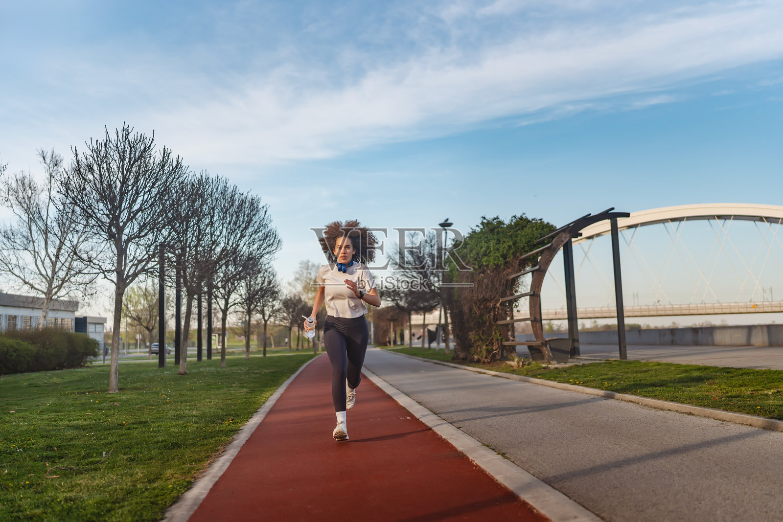 一位年轻女性在现代桥附近户外慢跑,穿着白色T恤、深色紧身裤,脖子上挂着耳机,手里拿着水瓶,神情专注。照片摄影图片