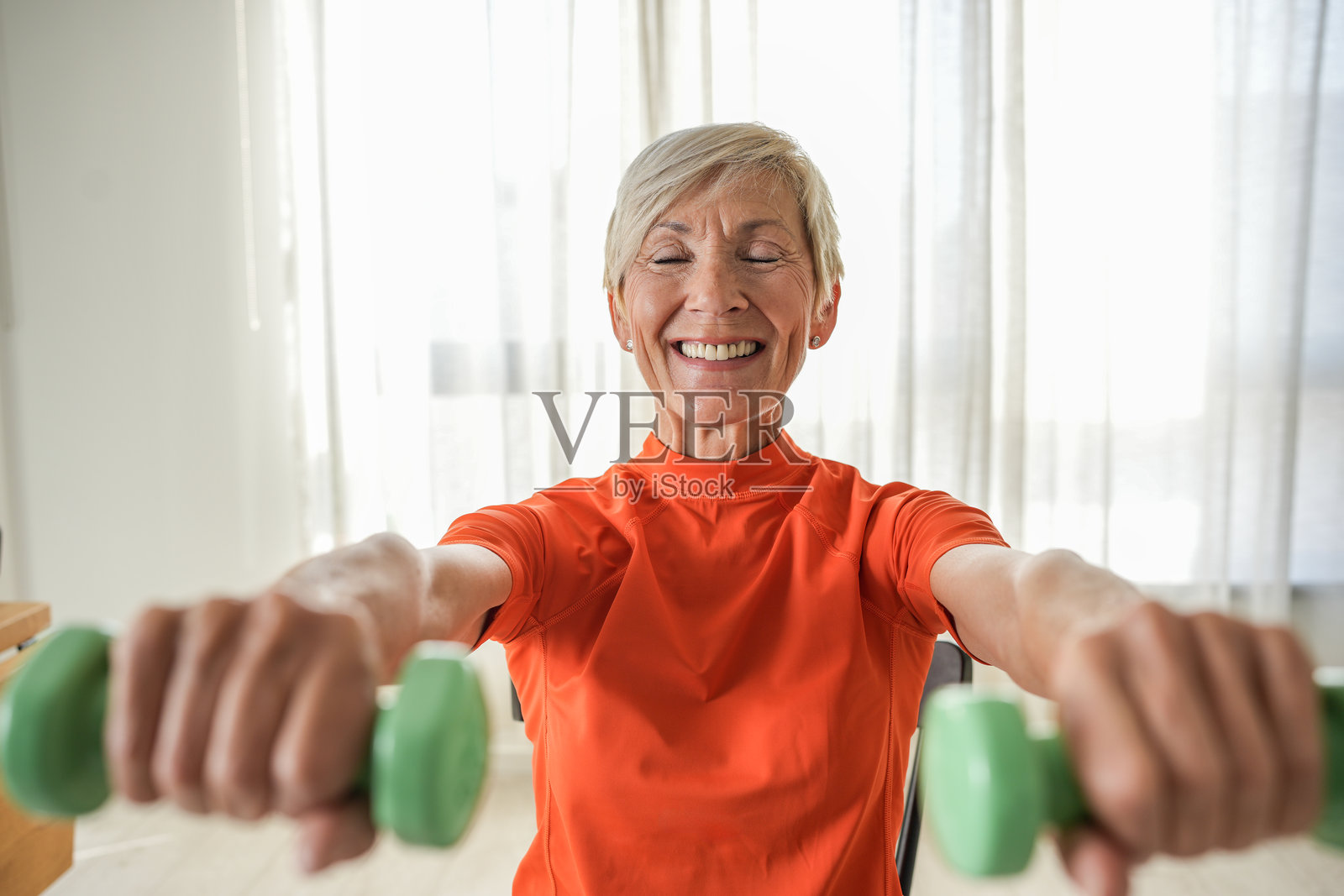 一位老年女性坐在椅子上,微笑着举起绿色哑铃。她在一个明亮的客厅里锻炼,周围有植物,展现出力量、健康和积极的生活方式。照片摄影图片