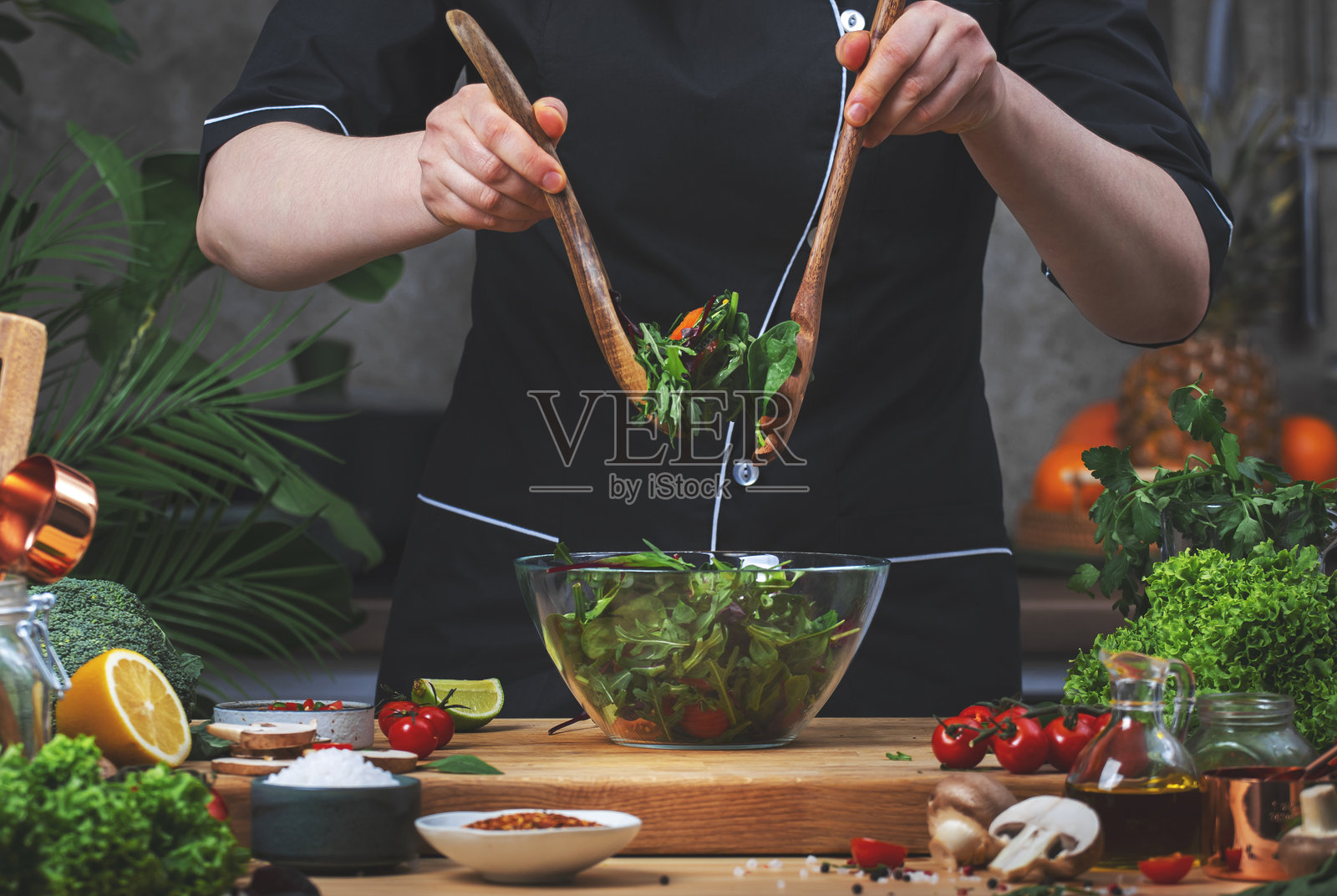 在厨房烹饪食物。厨师在木桌上用厨房用具、蔬菜、香草和配料搅拌碗中的绿色沙拉和芝麻菜。健康的素食饮食。照片摄影图片