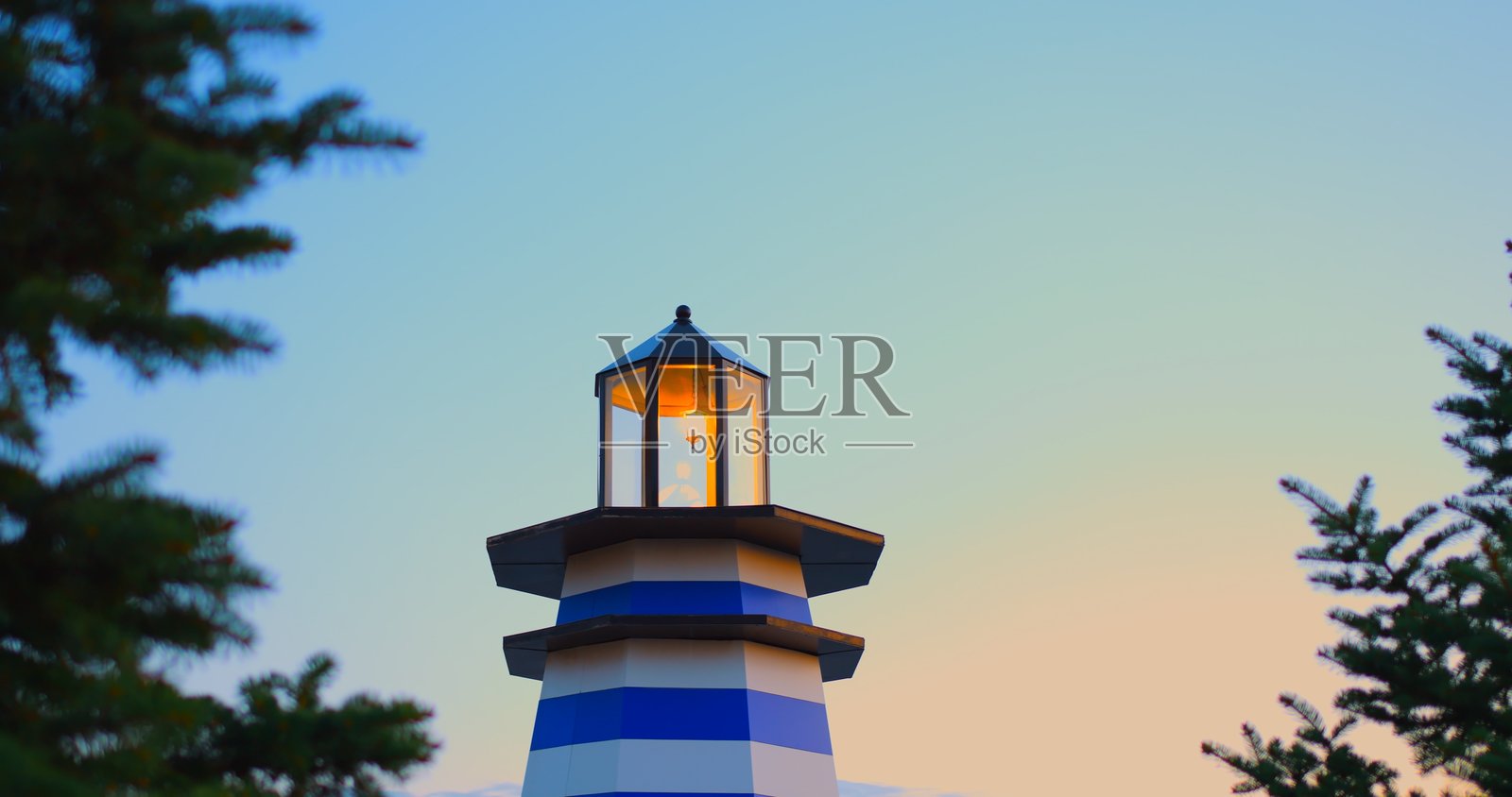 装饰性的灯塔,发出明亮的灯光,照亮蓝白相间的条纹,矗立在清澈的日落天空下,松树枝在金色的晚光中投下剪影。照片摄影图片