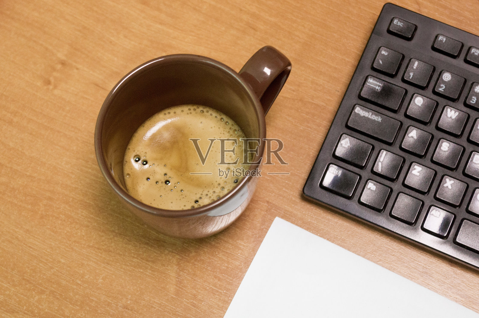 高角度拍摄的木桌,上面有一个棕色咖啡杯、黑框眼镜放在蓝色纸上,以及部分黑色键盘,展现出一种休闲现代的工作空间布置。照片摄影图片