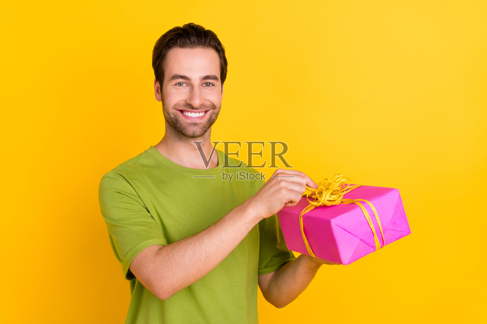 快乐的胡须千禧年男子手持礼物,穿着绿色T恤,背景为黄色。照片摄影图片