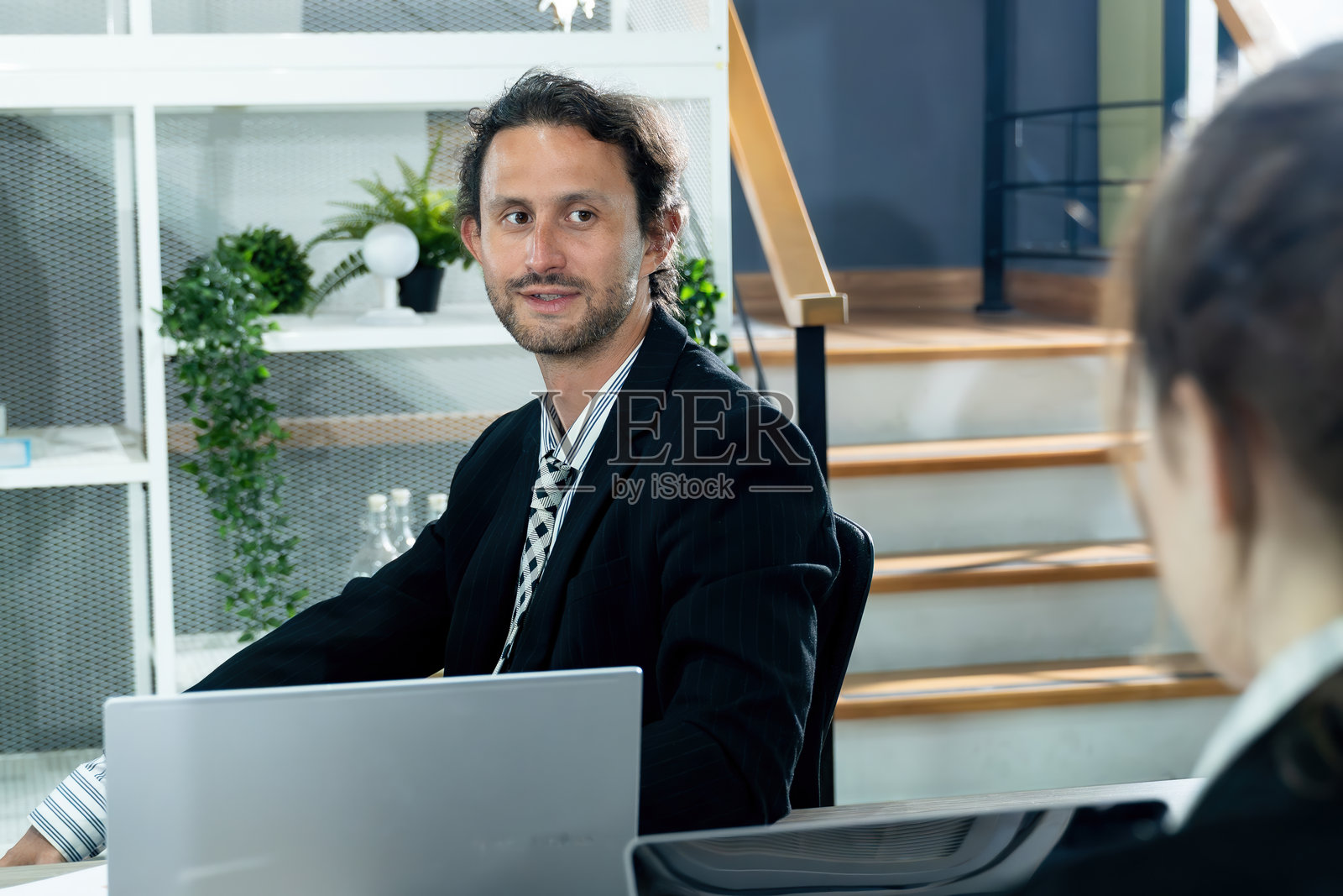 年轻的商务人士穿着西装,坐在办公室桌前,专注地与同事交谈,进行轻松的商务讨论,工作空间中的企业环境,多元文化团队合作照片摄影图片