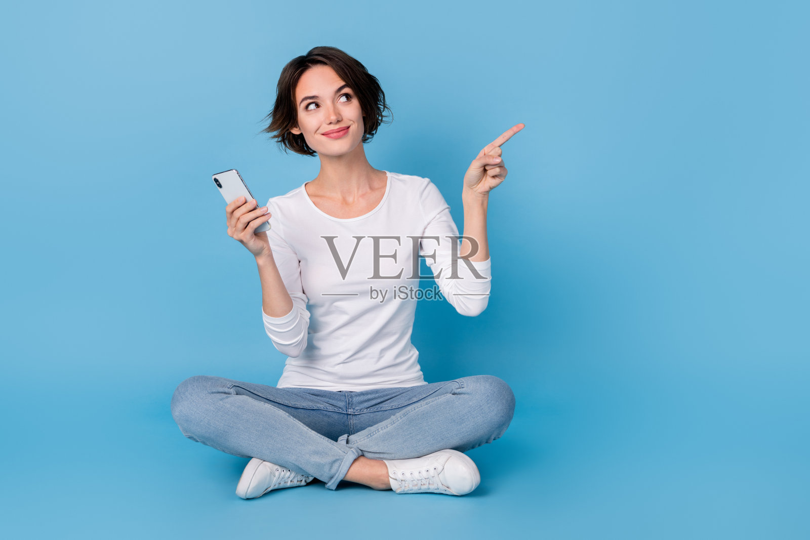 一位穿着白衬衫、双腿交叉、手持现代设备的搞笑女士,背景为空白的蓝色。照片摄影图片