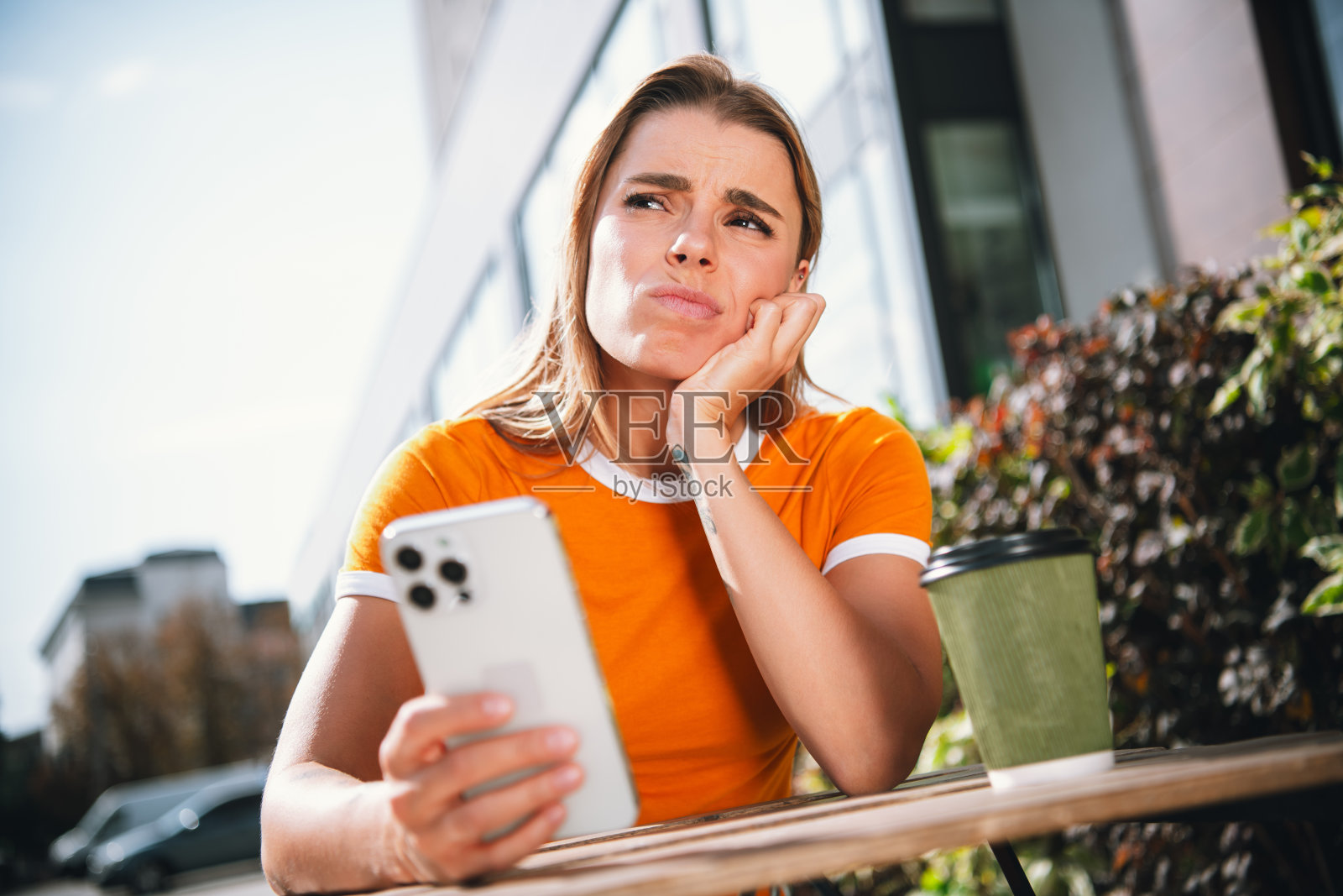 年轻女性穿着橙色T恤,在城市环境中户外拿着智能手机,神情若有所思,阳光明媚。照片摄影图片