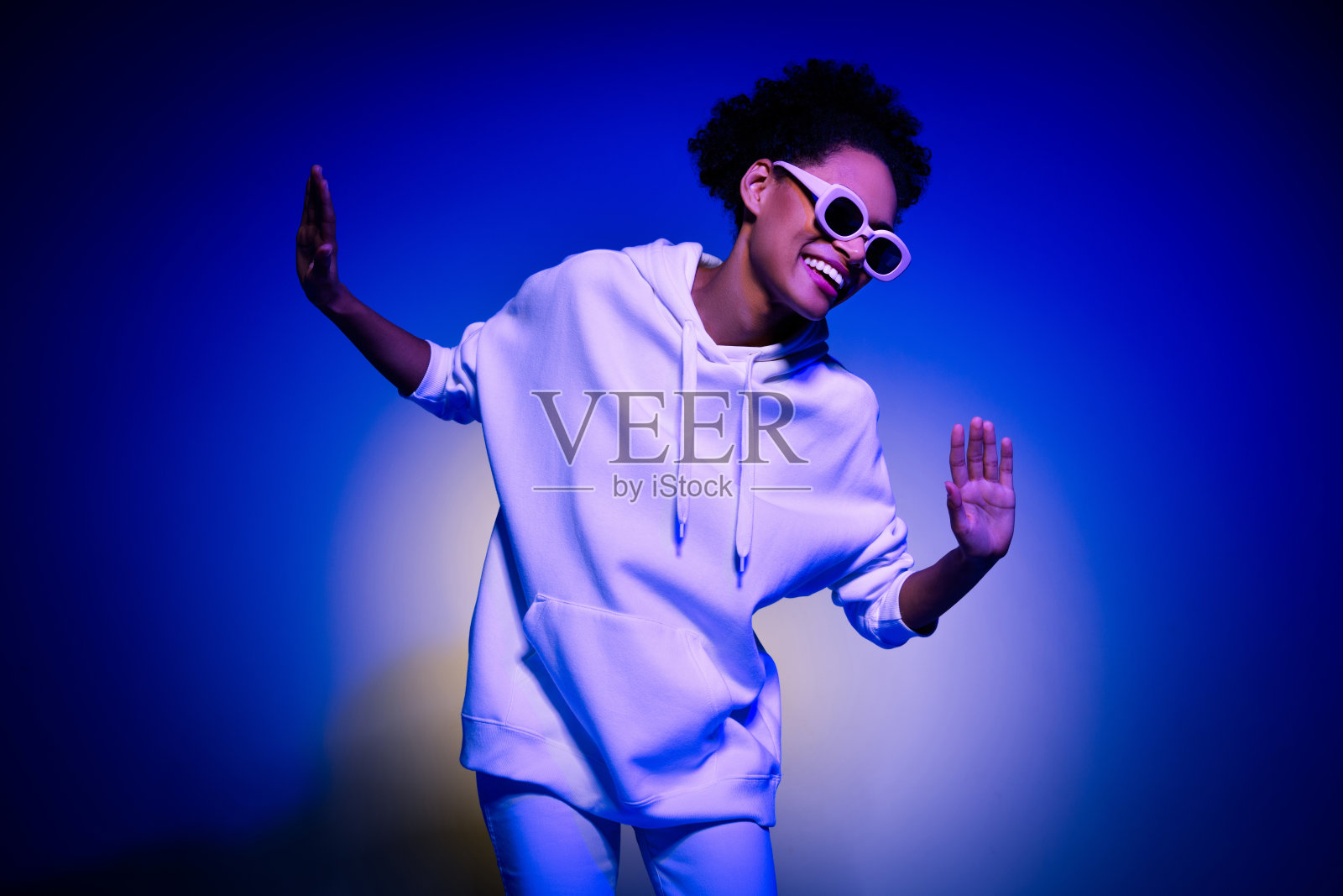 穿着卫衣、戴着深色眼镜的甜美漂亮女性在蓝色背景下微笑跳舞的照片照片摄影图片