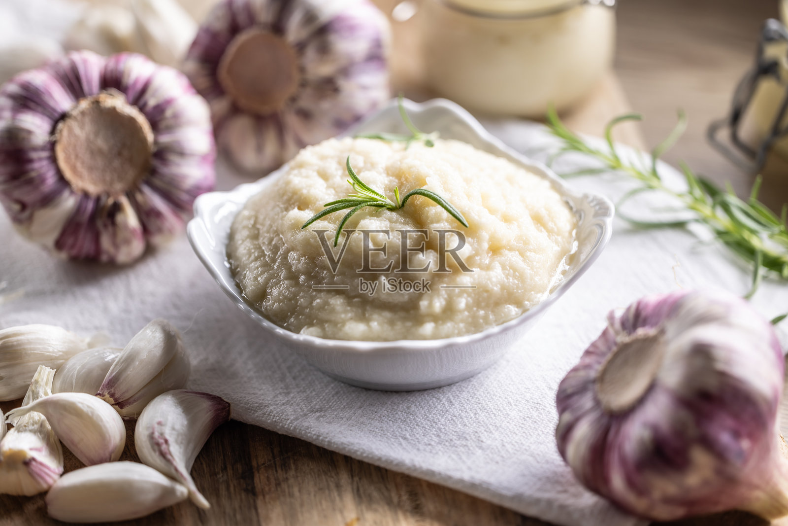 香气四溢的蒜蓉放在铺有乡村风格布料的碗中,旁边有蒜头、剥好的蒜瓣和迷迭香。照片摄影图片