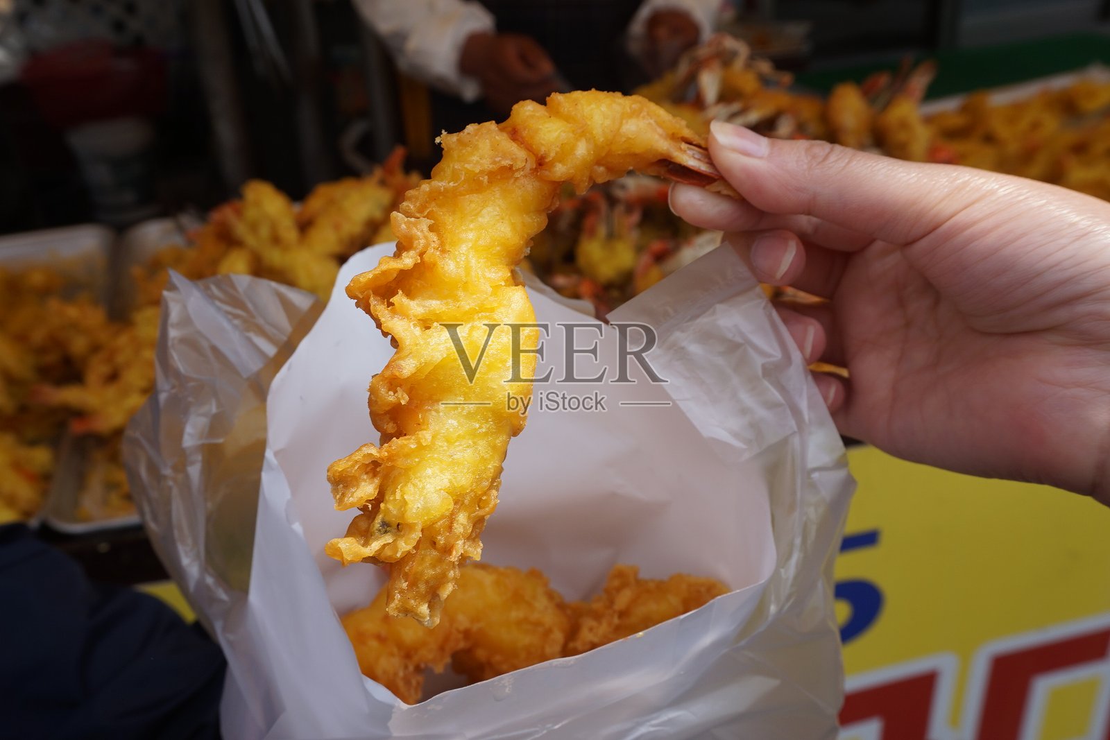 一只手将酥脆金黄的炸虾放入白色塑料袋中,背景中还有额外的炸虾托盘,突显新鲜街头食品的制作过程。照片摄影图片