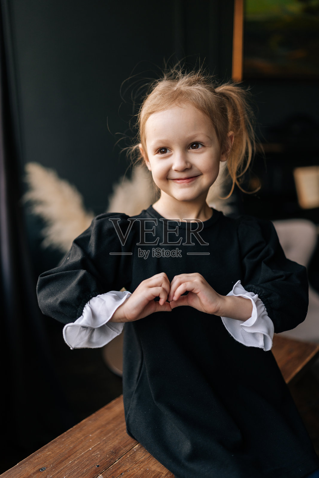 可爱的小女孩,金色头发,穿着黑色裙子,坐在木凳上用手做出心形,展现出在温馨室内环境中的爱与亲情。照片摄影图片