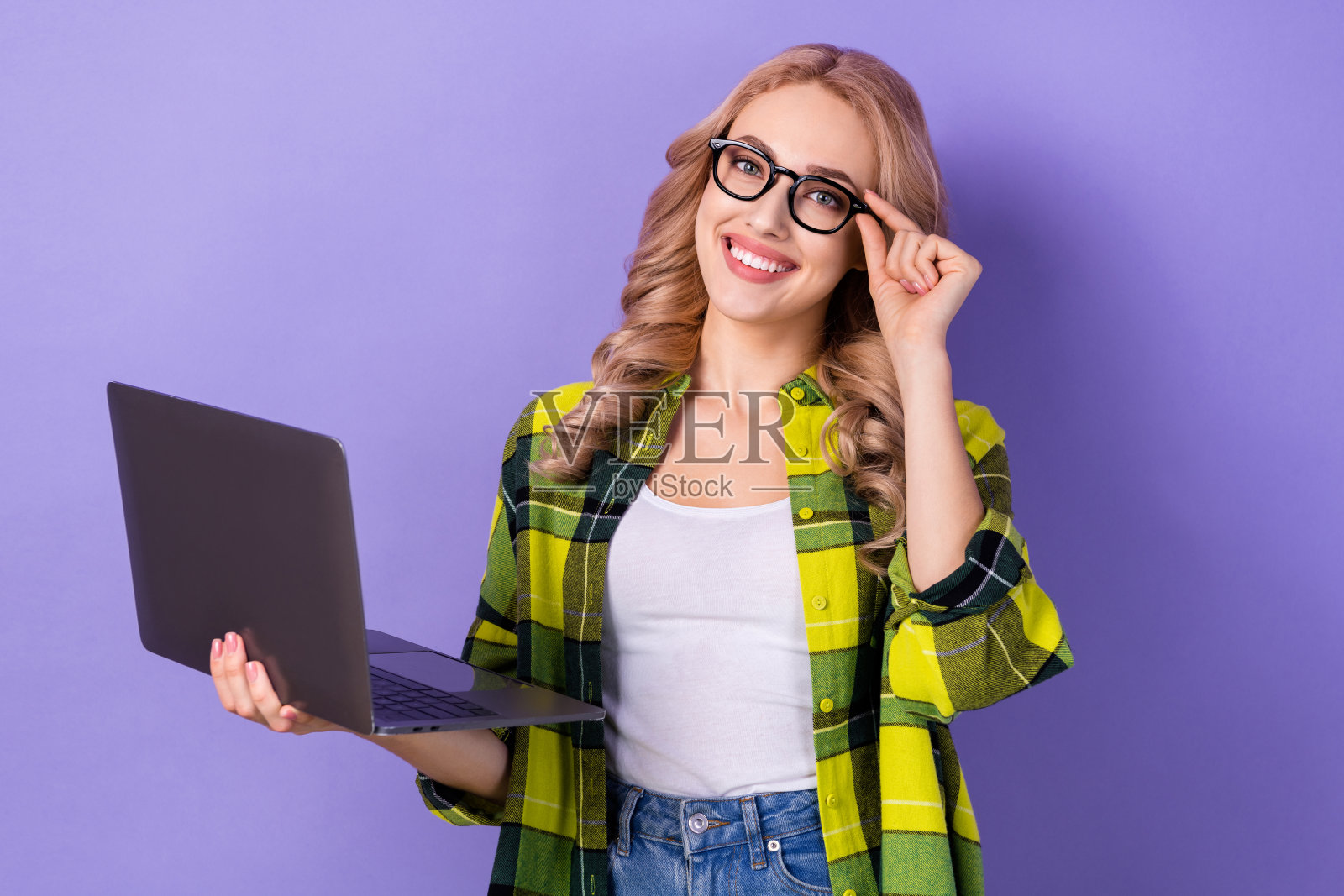 漂亮积极的女孩手臂触碰眼镜,露出灿烂的笑容,手持无线笔记本电脑,背景为紫色。照片摄影图片
