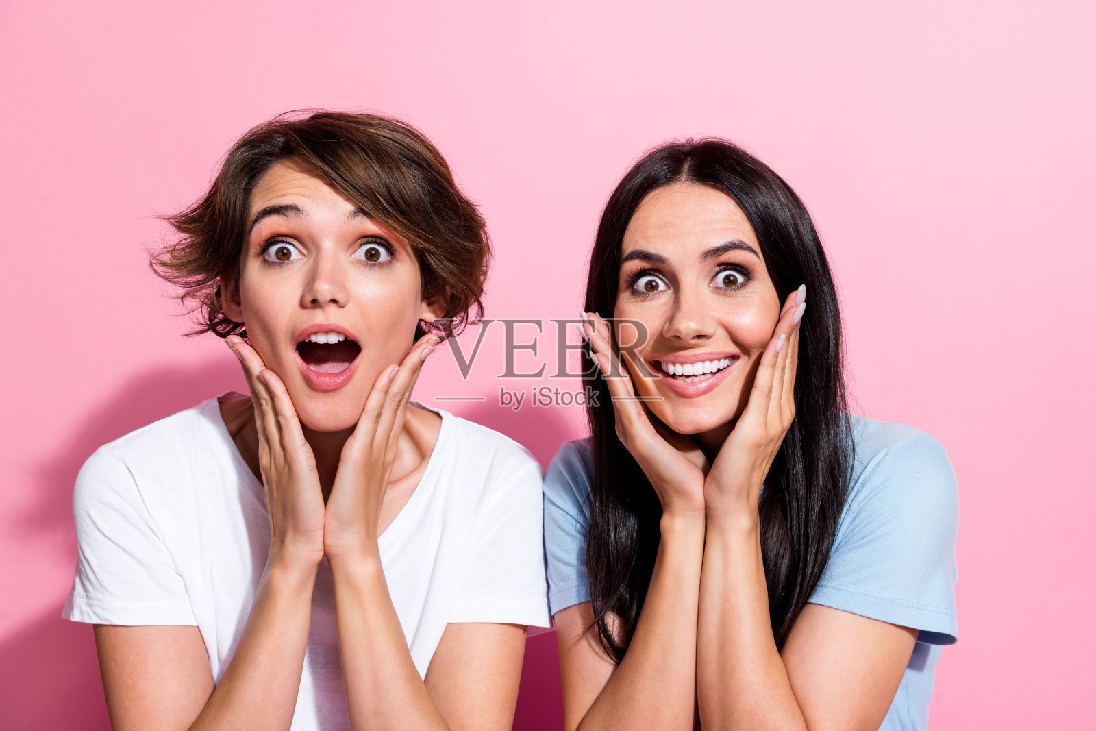 两个女孩的特写肖像,面露惊讶,双手捂脸,惊叫“哇哦”,在粉色背景下表现出意外的反应。照片摄影图片