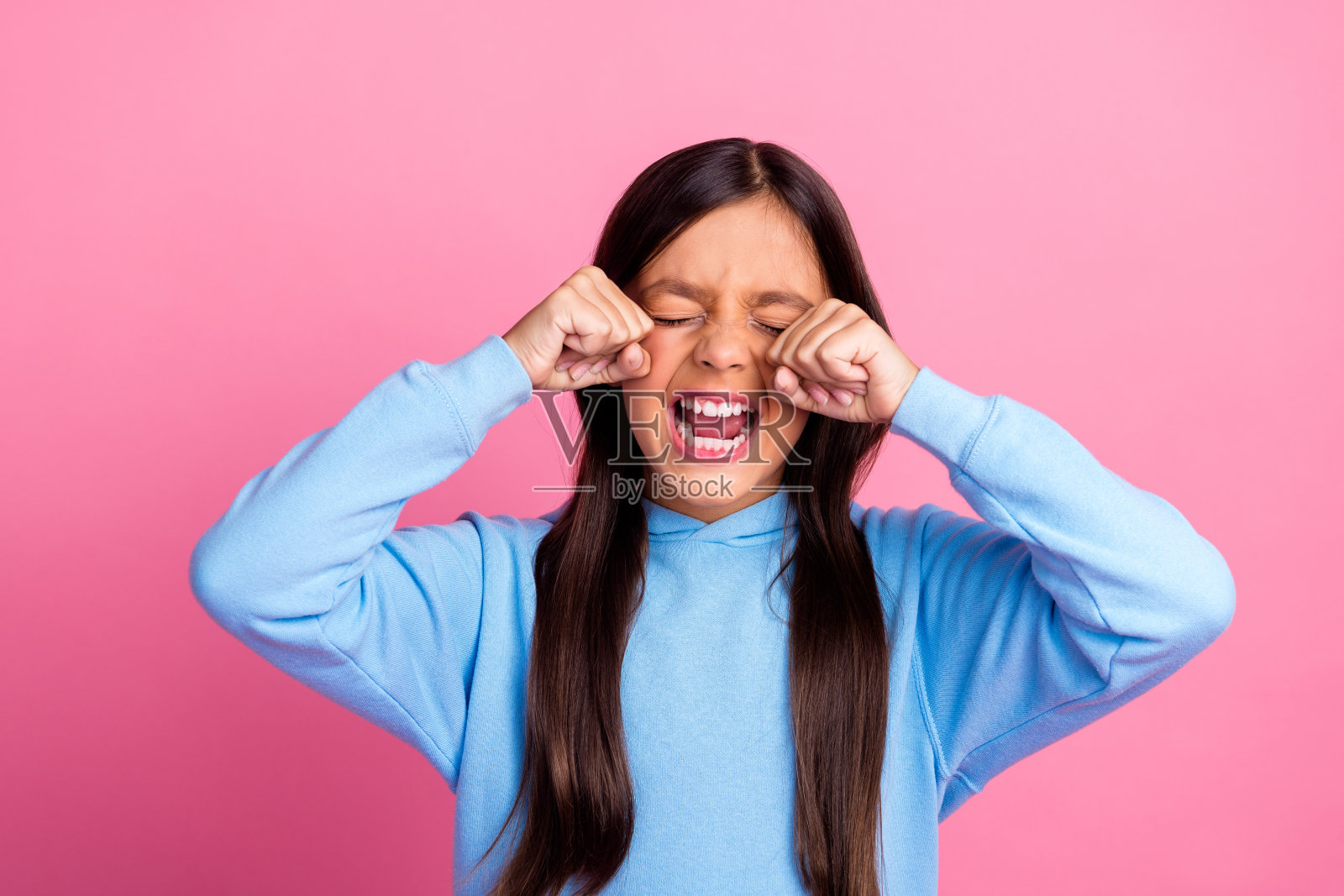 穿着蓝色卫衣的女孩在粉色背景前揉眼睛,表现出可爱的情感和生动的手势。照片摄影图片