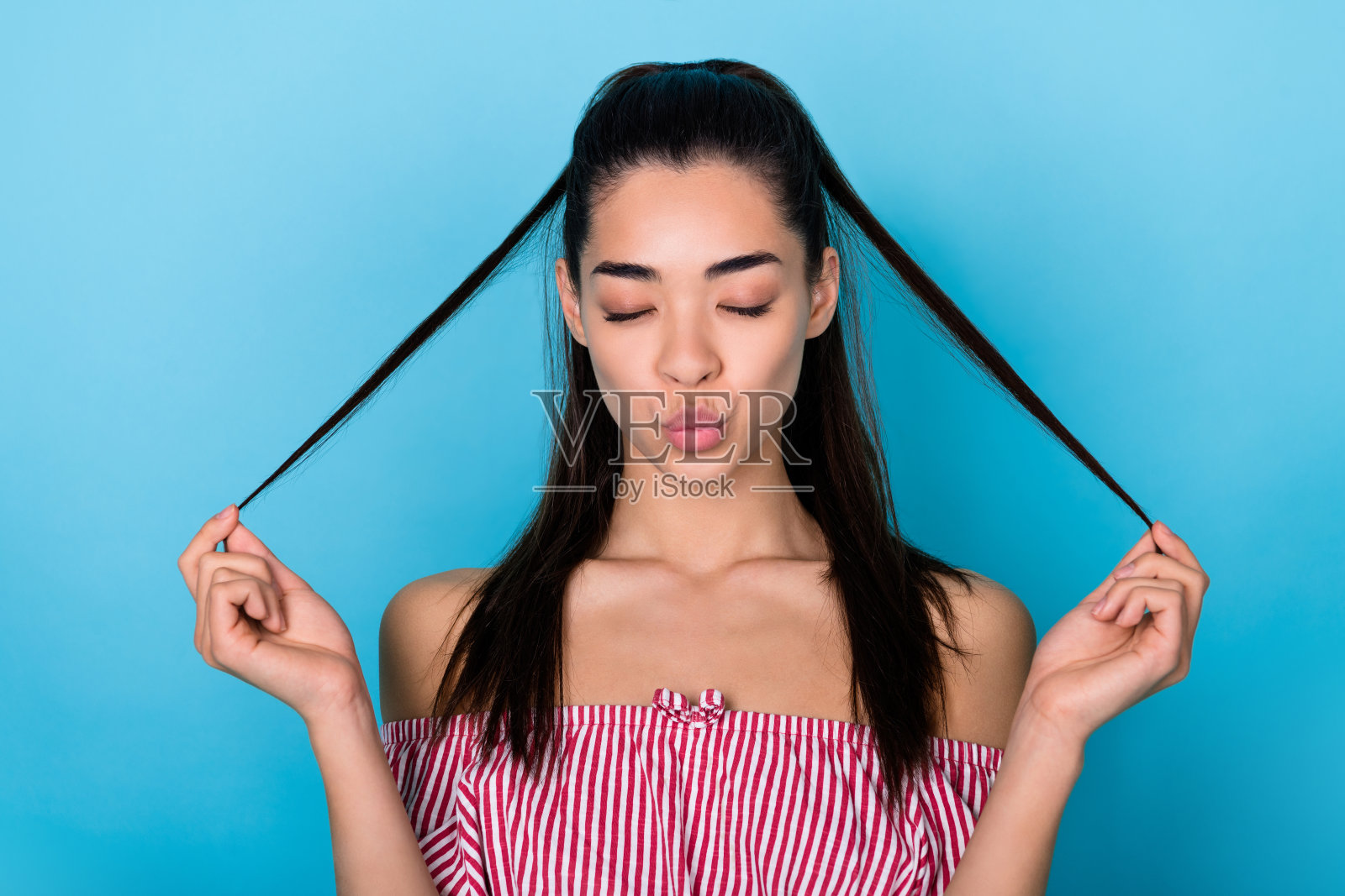 一位时尚女性的肖像,撅嘴亲吻,丰盈的嘴唇,棕色的马尾辫,穿着红色上衣,闭着眼睛,背景为蓝色。照片摄影图片