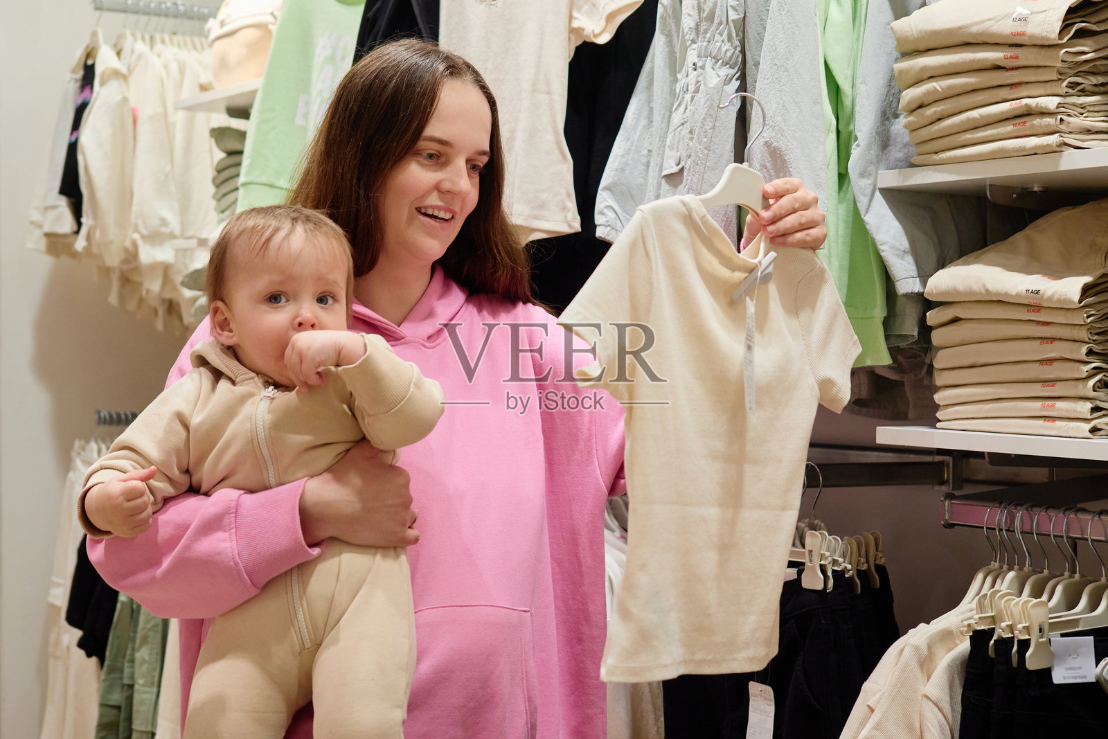 一位母亲抱着她的宝宝,在零售店里挑选衣服。她正在查看架子上展示的一件可爱衬衫,展现出愉快的购物体验。照片摄影图片