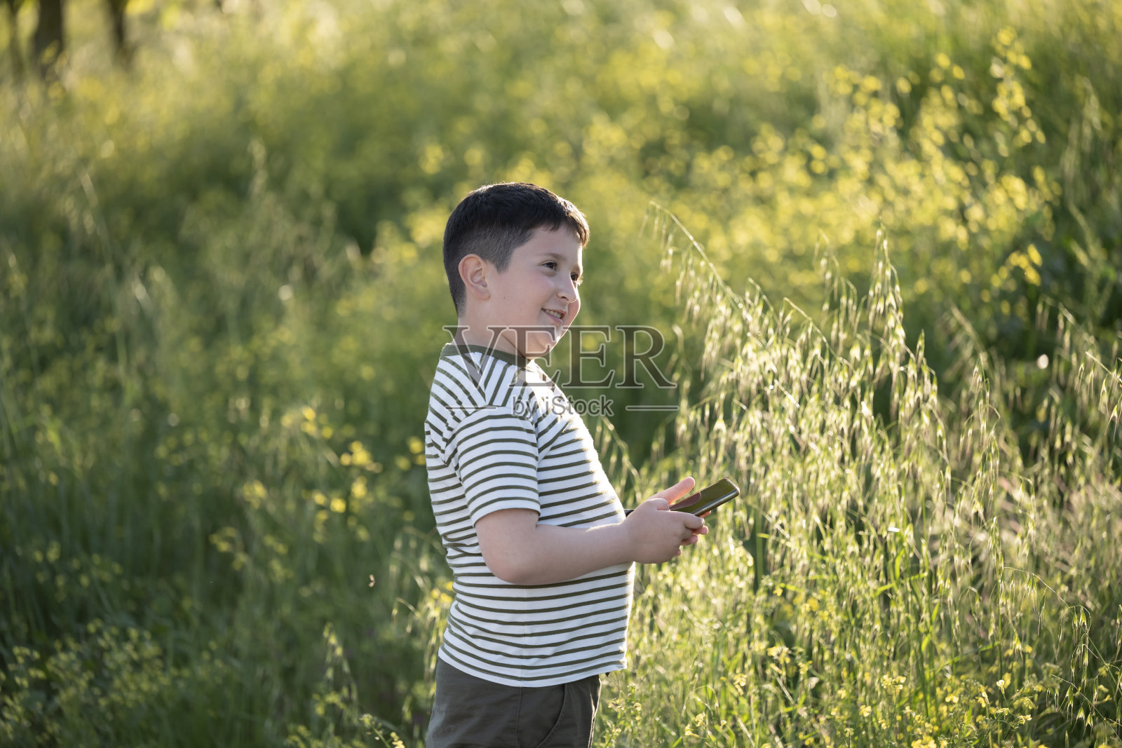 一位吸引人的少年男孩手持智能手机在绿叶背景下自拍。利用智能手机技术在户外拍照。照片摄影图片