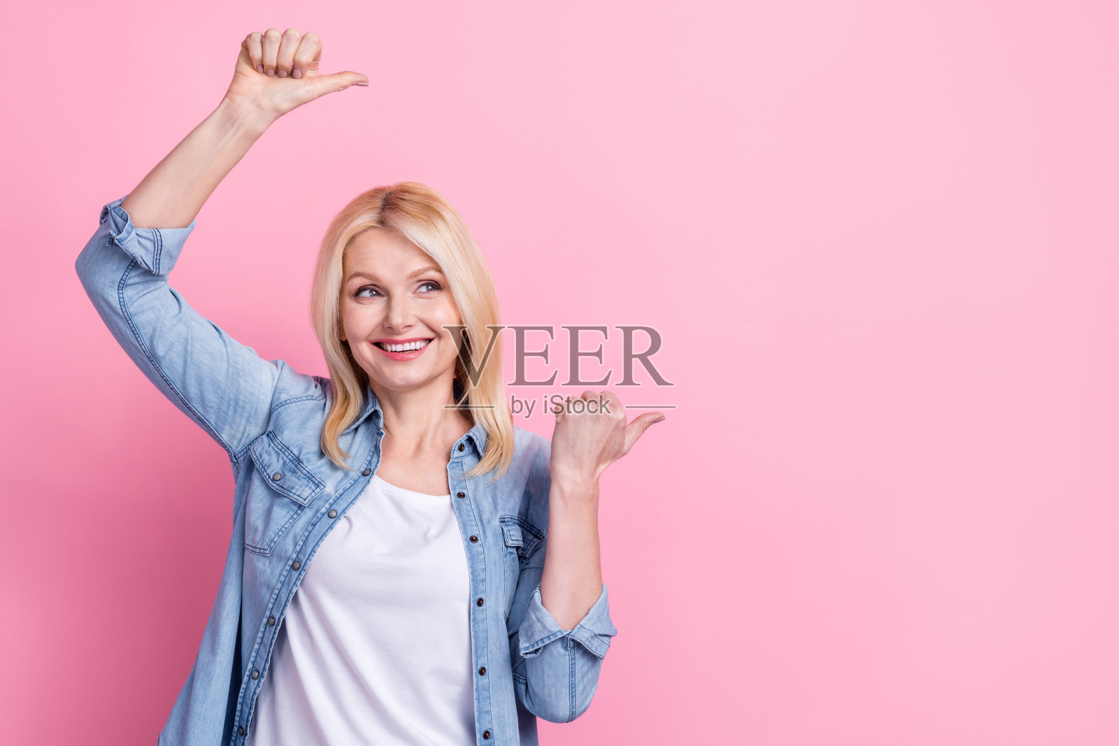 可爱的退休女士竖起大拇指,展示空白空间,穿着牛仔裤和衬衫,背景为粉色。照片摄影图片