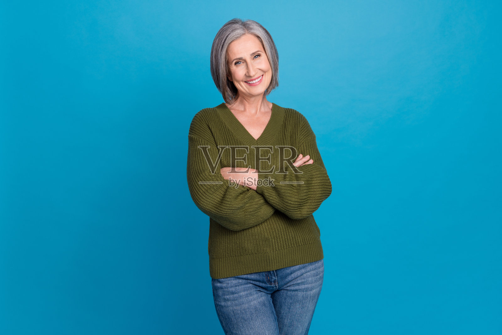 微笑着露齿而笑的快乐人士,灰色发型,穿着针织毛衣,双臂交叉,背景为蓝色。照片摄影图片