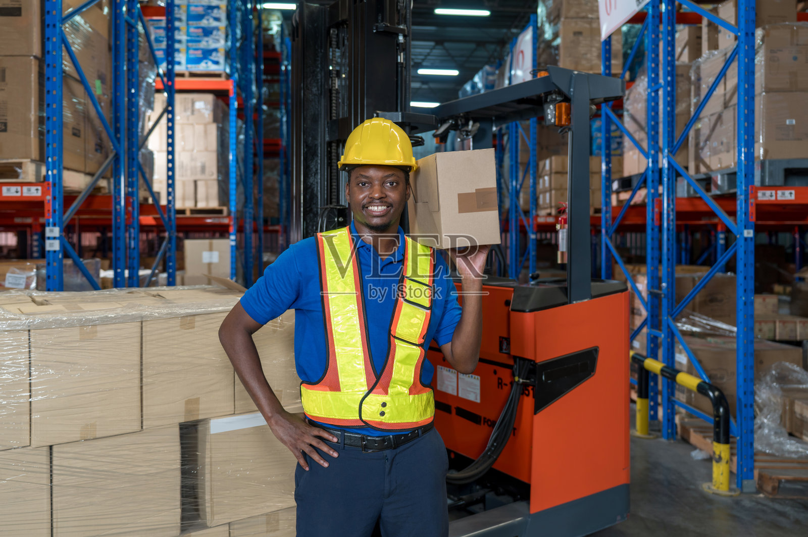 一名穿着安全装备的男子在一个井然有序的仓库中手持一个大纸箱。货架上摆满了各种产品,显示出繁忙的工作环境。照片摄影图片
