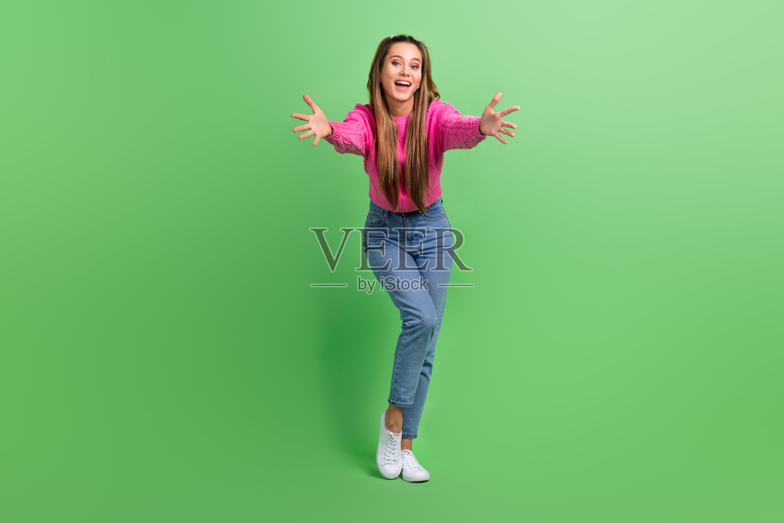 全身照,愉快积极的女孩穿着粉色毛衣和牛仔裤,伸出双手想要拥抱你,背景为绿色。照片摄影图片