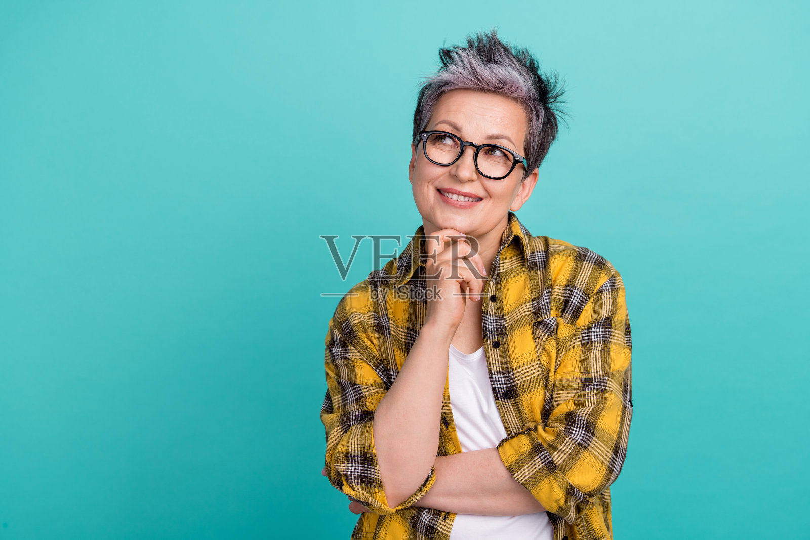 穿着时尚黄色格子衣服的迷人退休女性,手托下巴,目光凝视空旷的空间,背景为青色。照片摄影图片