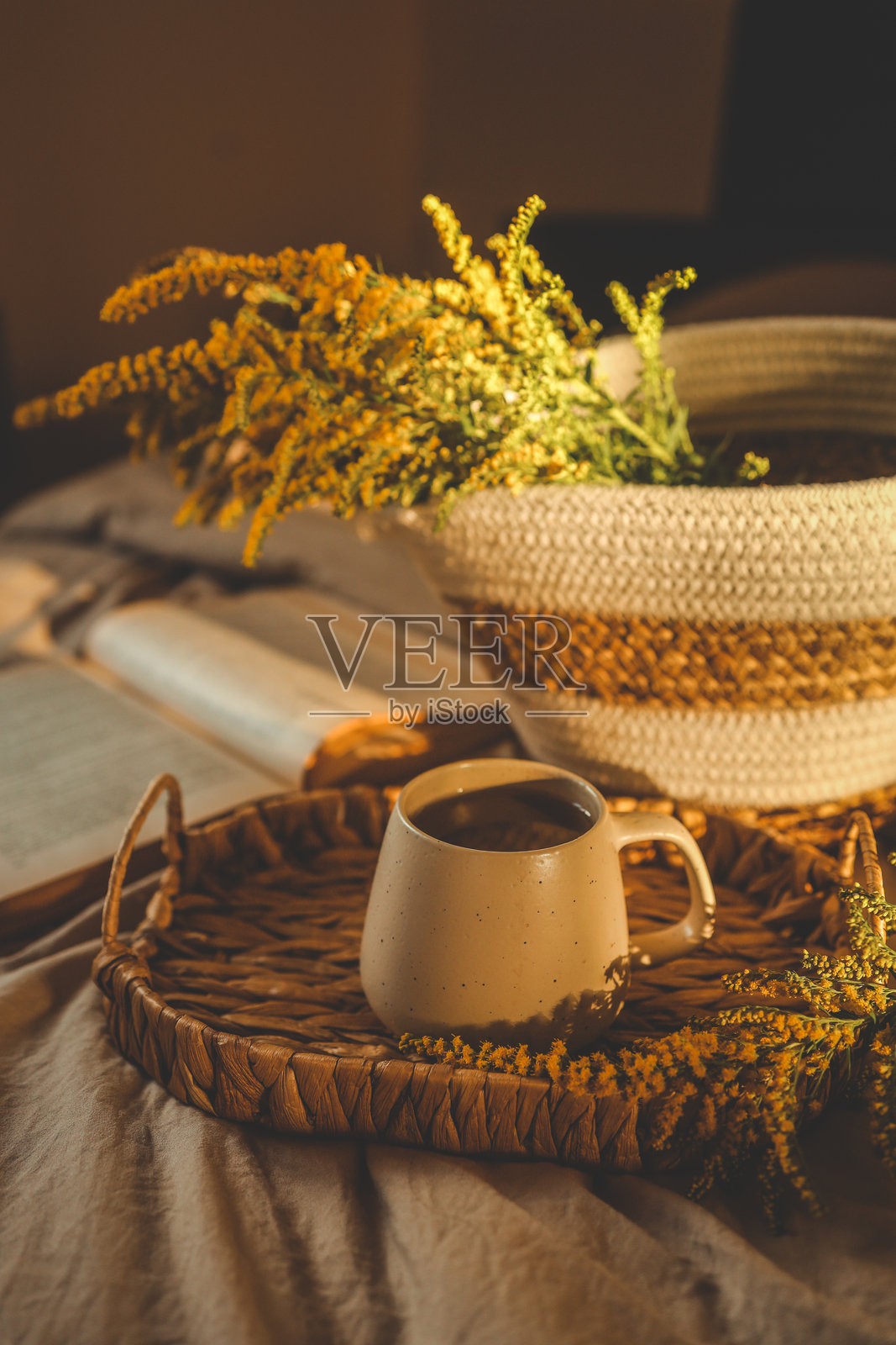 温暖的早晨阳光下,柔软的亚麻床上摆放着一个陶瓷咖啡杯、一个装有黄色野花的编织篮子和一本打开的书,营造出舒适的场景。照片摄影图片