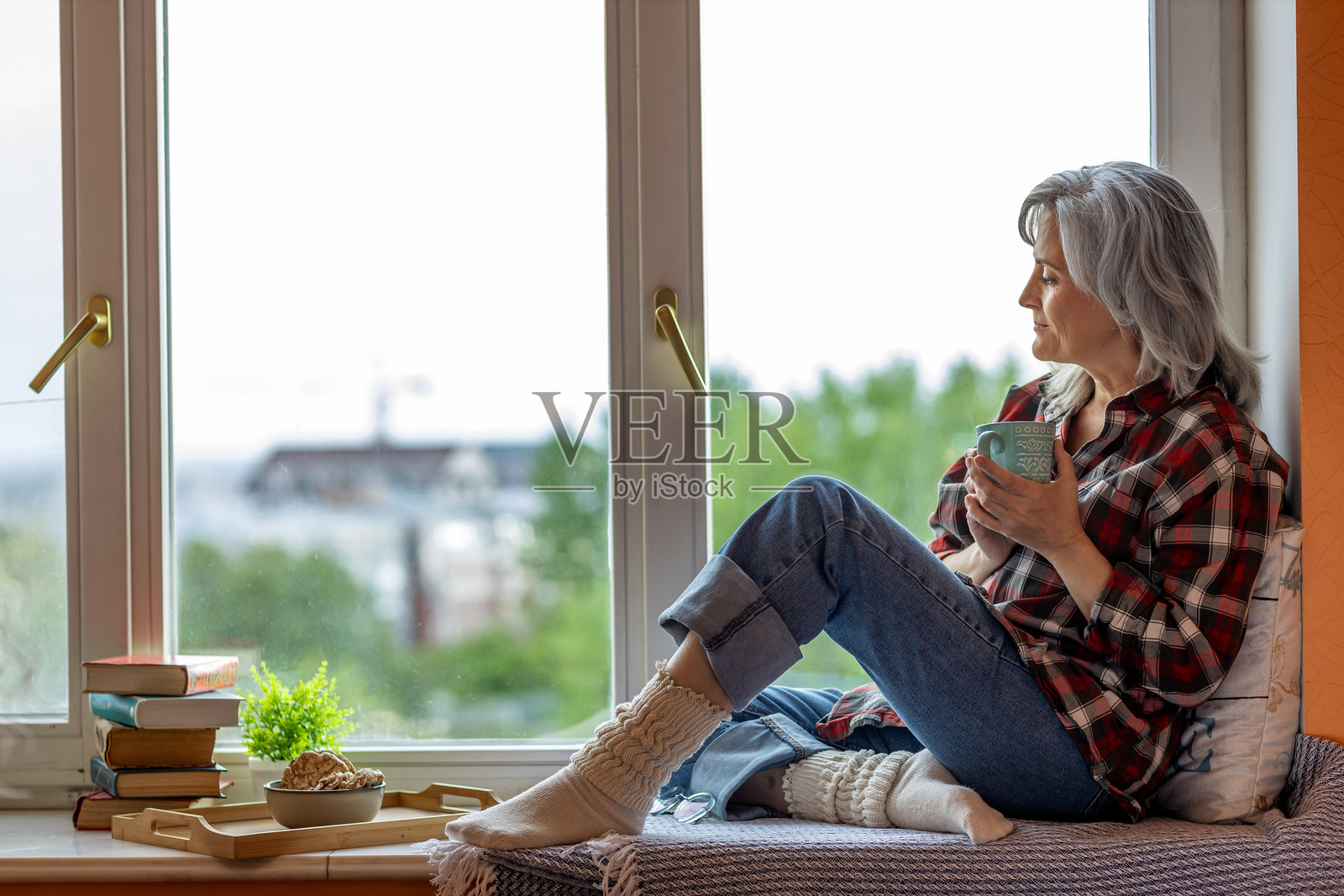 一位老妇人坐在窗台上,手里拿着茶和书,若有所思地望着窗外。照片摄影图片