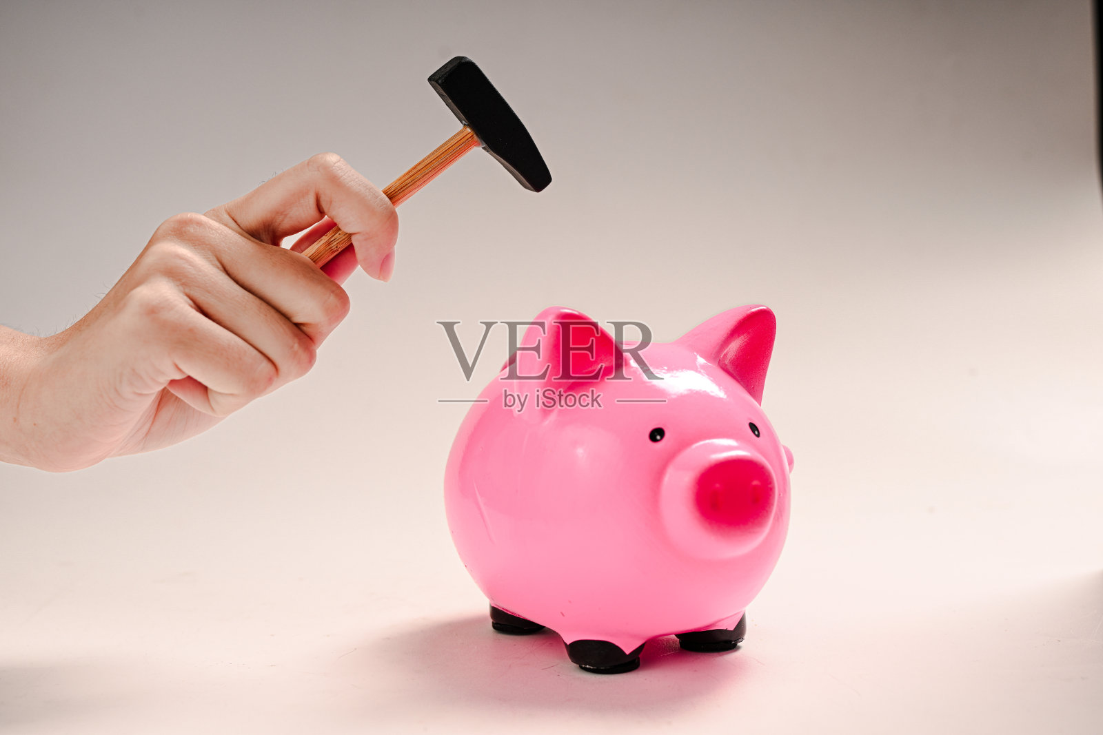 一只手握着一把小锤子,举在光滑的粉色储蓄罐上方,背景为奶油色,象征着打破储蓄或获取存储的钱。照片摄影图片