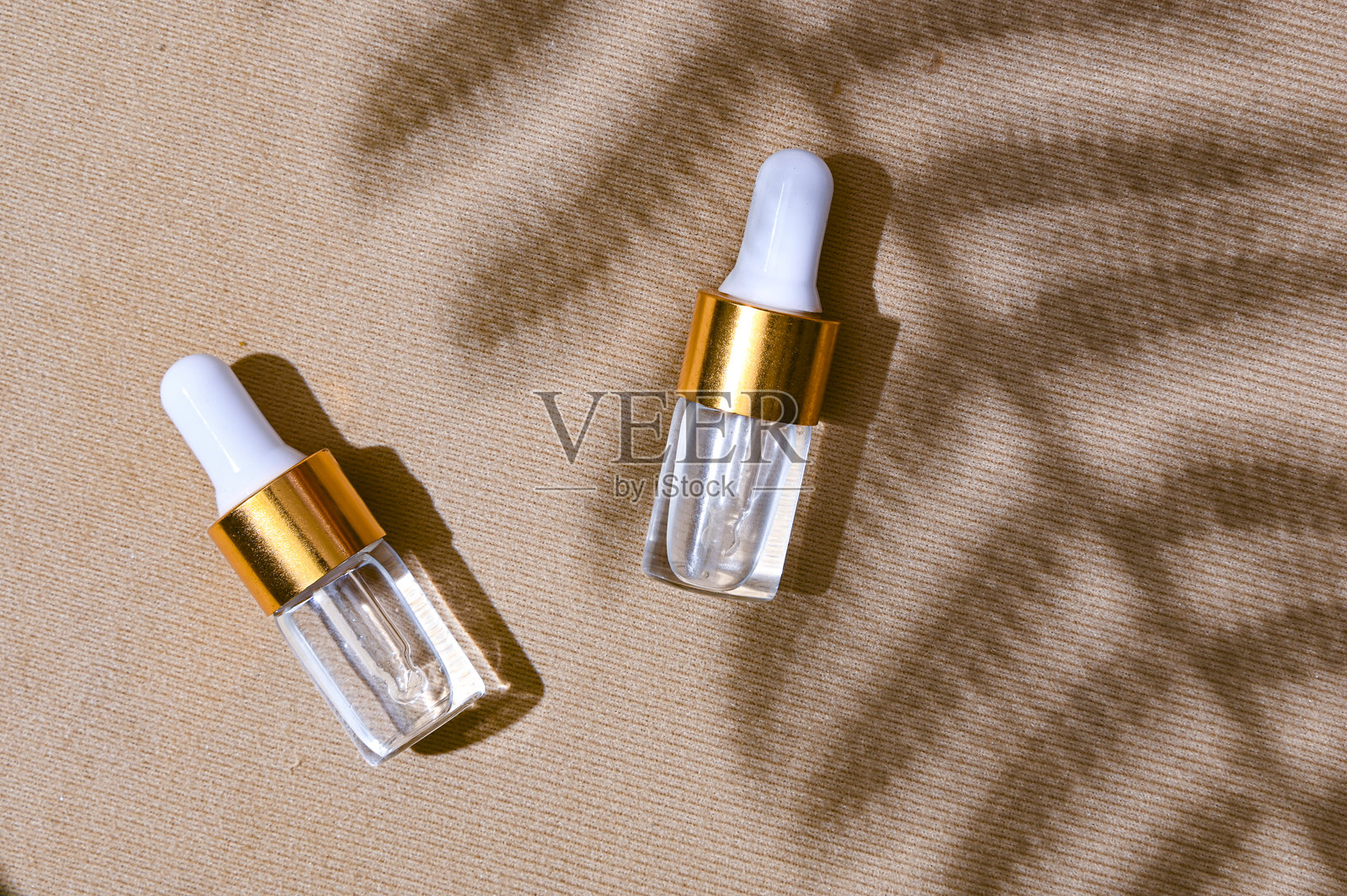 两个透明的滴管精华瓶,配有白色瓶盖和金色瓶颈,放置在有质感的米色背景上,背景中有柔和的叶子阴影。自然护肤品牌概念,散发夏日气息。照片摄影图片