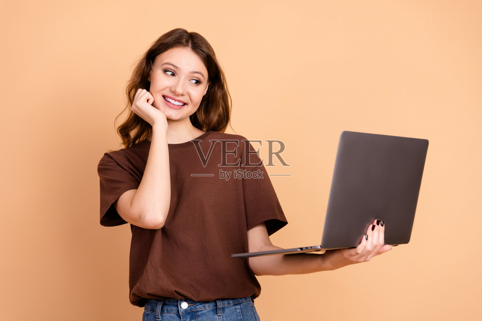 穿着棕色T恤的年轻女性微笑着,手里拿着一台笔记本电脑,背景是米色的。照片摄影图片