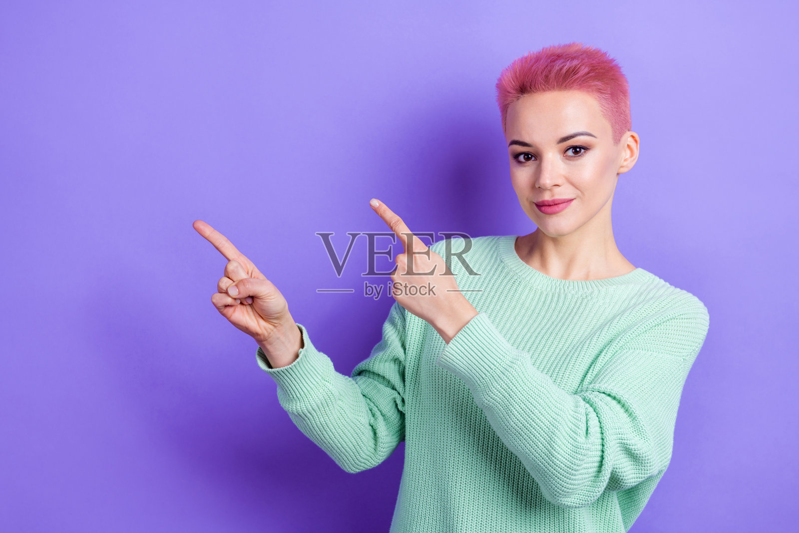 穿着青绿色毛衣的友好积极的人用手指指向空白空间,背景为紫色。照片摄影图片