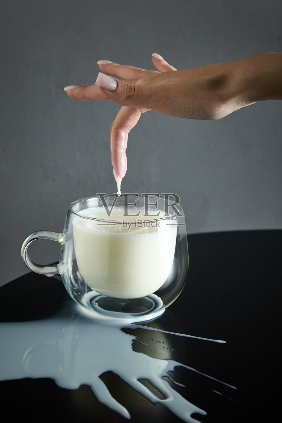 精致的手指浸入溢满的牛奶杯中,液体溢出在黑色表面上,饮品美学广告的概念照片摄影图片
