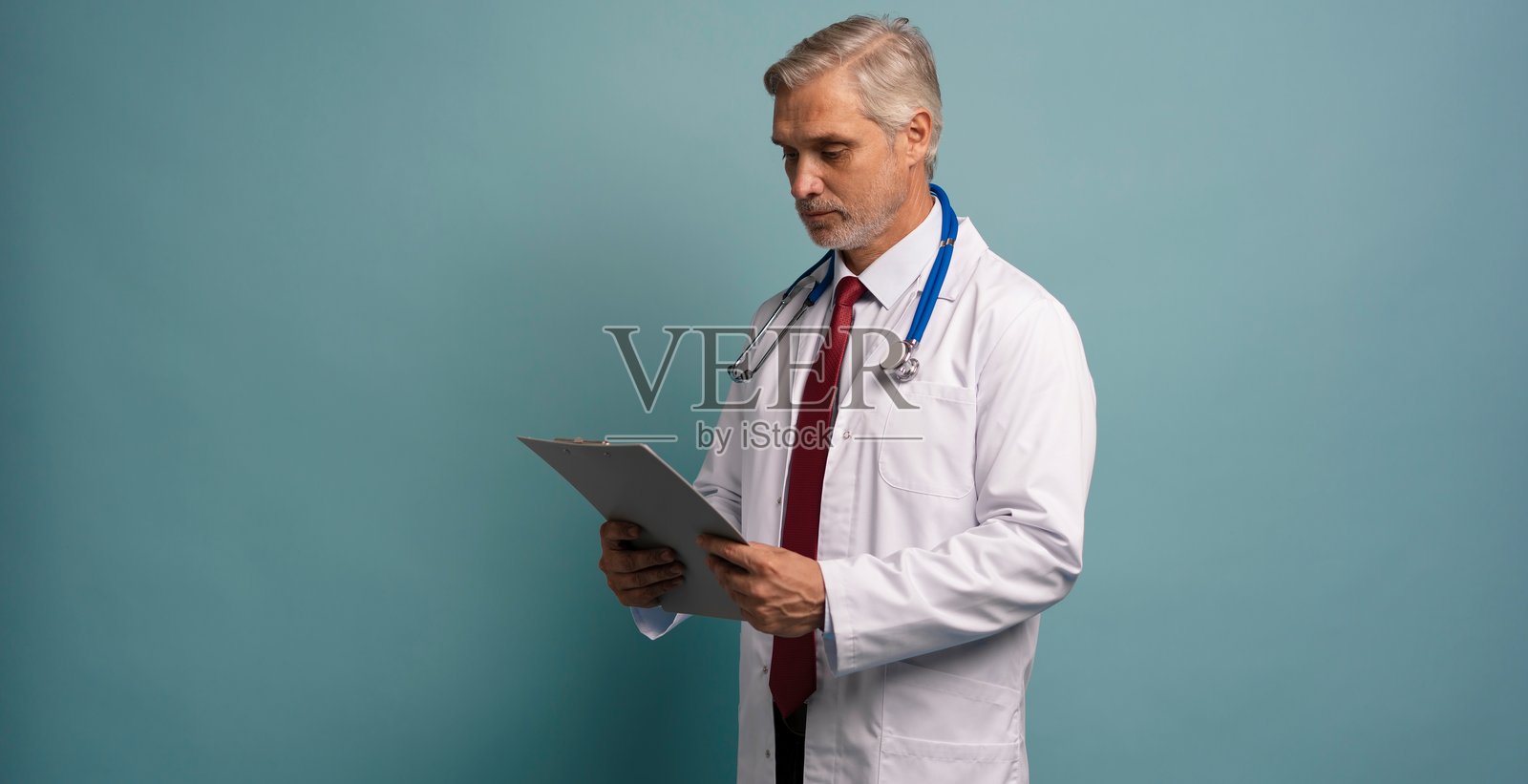 成熟的医疗专家,佩戴听诊器,微笑着看向镜头,手中拿着带有笔记的文件。照片摄影图片