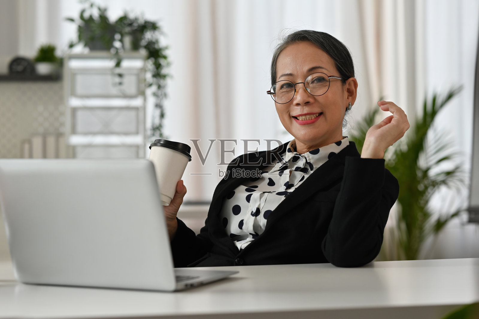 微笑的老年女商人在桌子旁坐着,戴着眼镜,手里拿着咖啡杯。她在明亮、现代的办公室环境中显得放松而自信。照片摄影图片