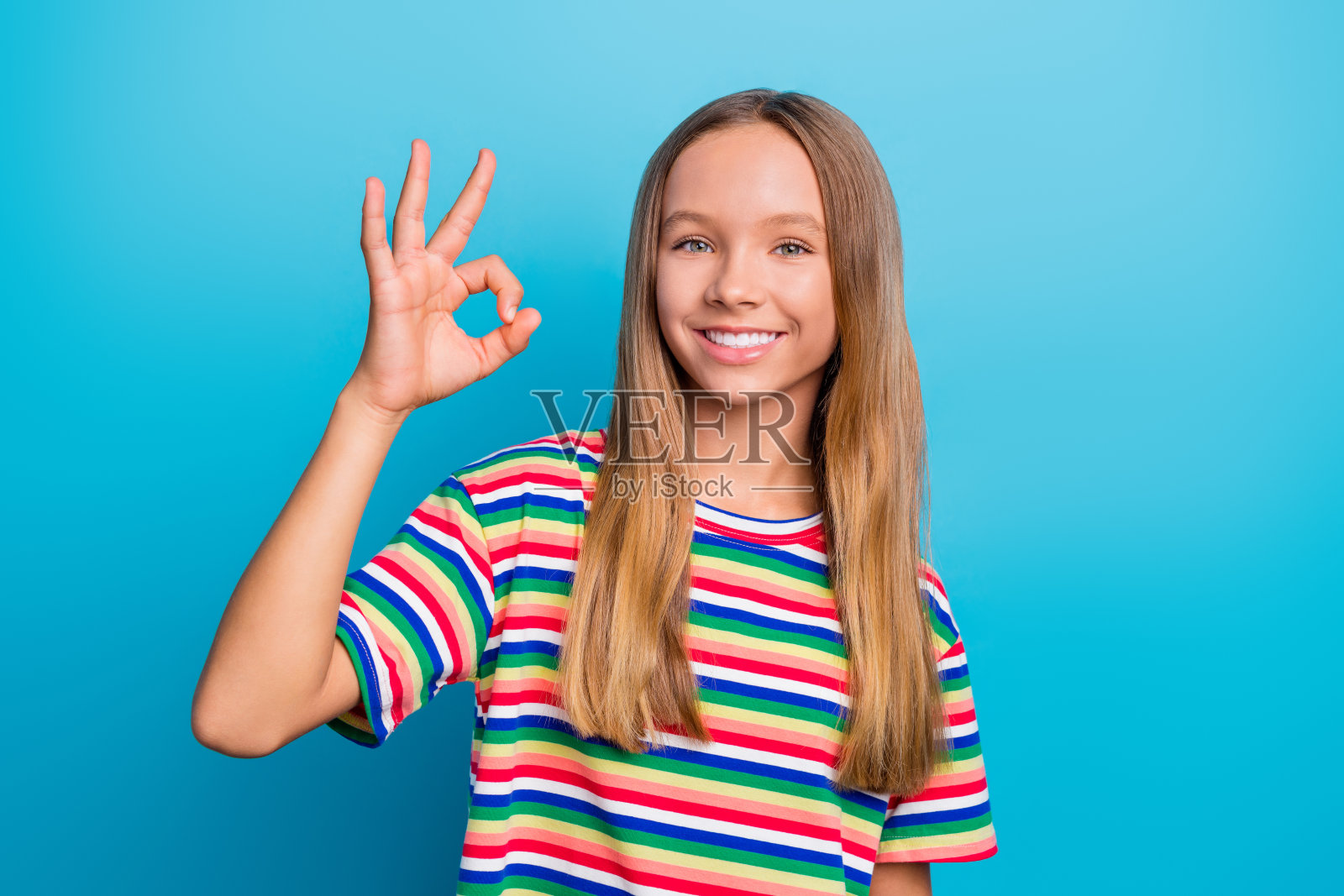 乐观的女学生肖像,长发,穿着彩色T恤,展示“好样的”手势,背景为蓝色。照片摄影图片