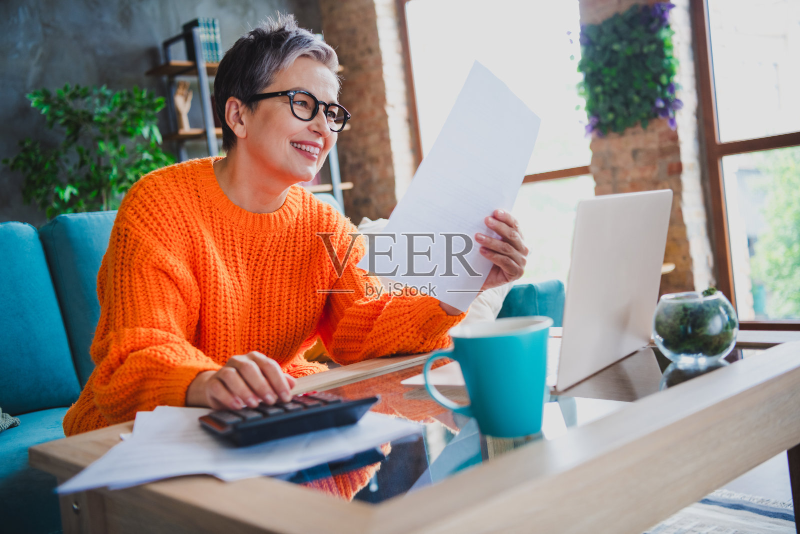 迷人的积极老年女士穿着橙色毛衣,在公寓室内用现代设备数记录。照片摄影图片