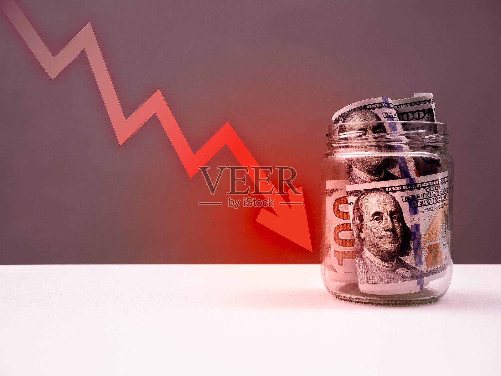 红色箭头射向玻璃罐内的美元钞票,罐子放在白色桌子上,背景为灰色。股市崩盘,美元贬值,商业金融,钱财概念。照片摄影图片