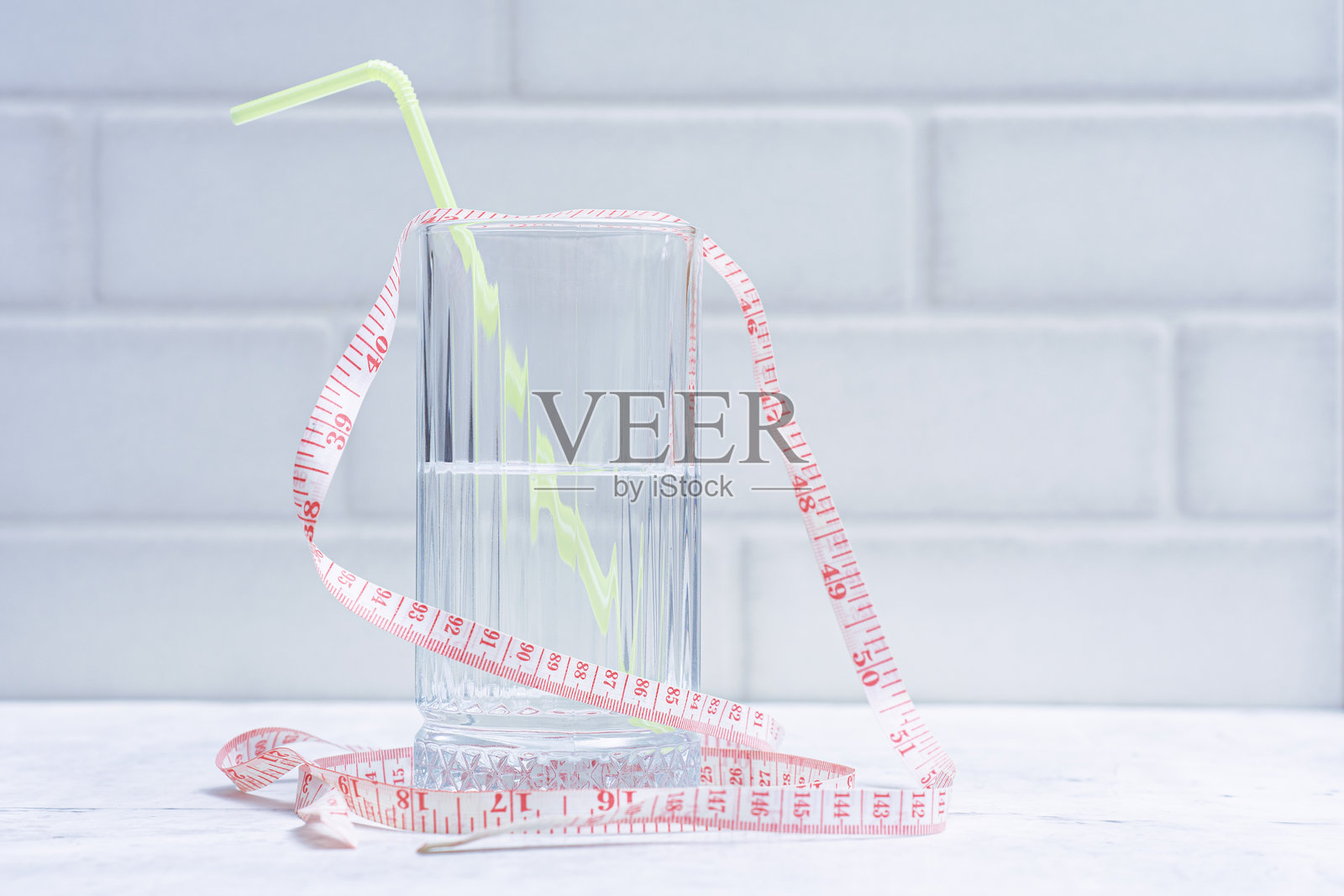 清水饮料杯,配有黄色吸管和粉色卷尺,背景为浅色。极简主义概念,适用于减肥、饮食和健康生活方式。照片摄影图片