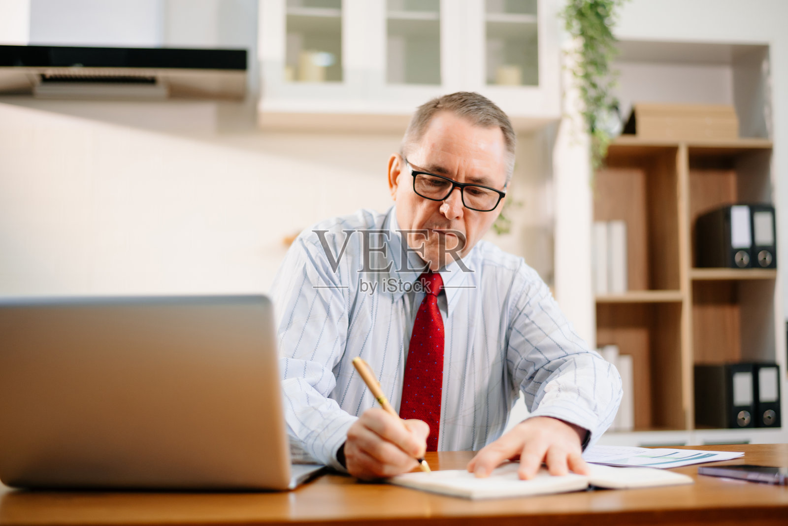 身穿红色领带的资深商人在现代厨房家庭办公室的桌子上使用笔记本电脑。远程工作,企业氛围。照片摄影图片