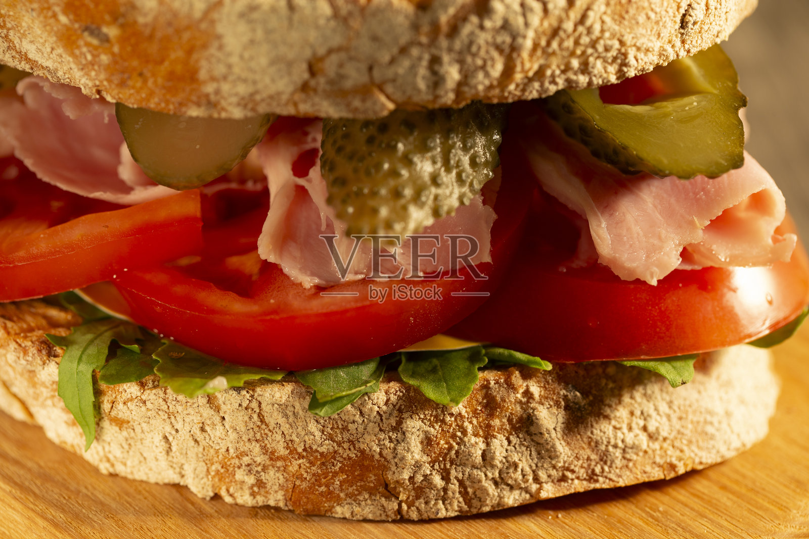 美味三明治特写,夹有火腿、萨拉米香肠、腌黄瓜和新鲜蔬菜,放在乡村风格的木砧板上,背景为木桌。照片摄影图片