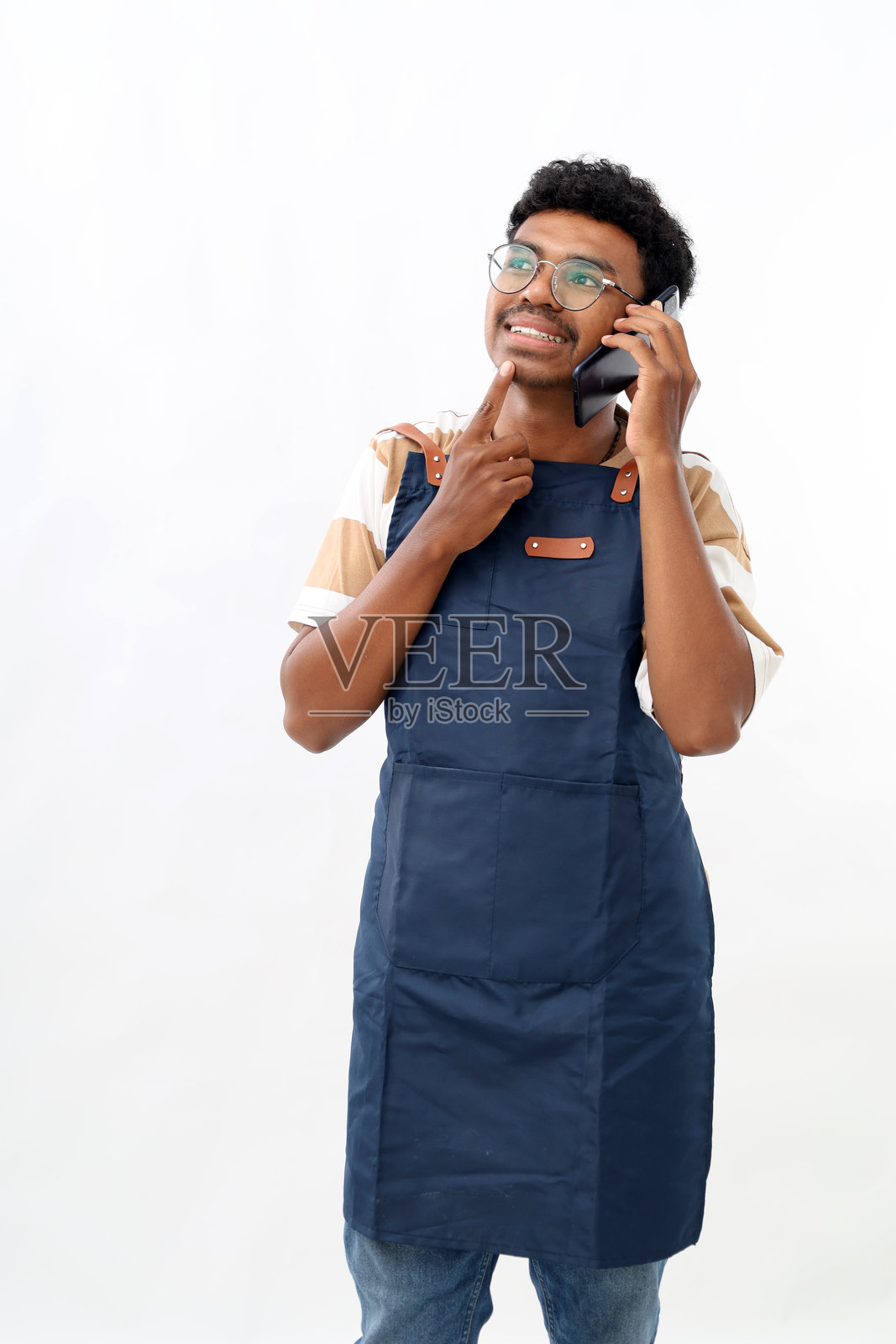 年轻有趣的亚洲男咖啡师穿着蓝色围裙,在咖啡店工作,手持手机与人交谈,托着下巴,目光投向一旁,背景为白色。小型创业公司。照片摄影图片