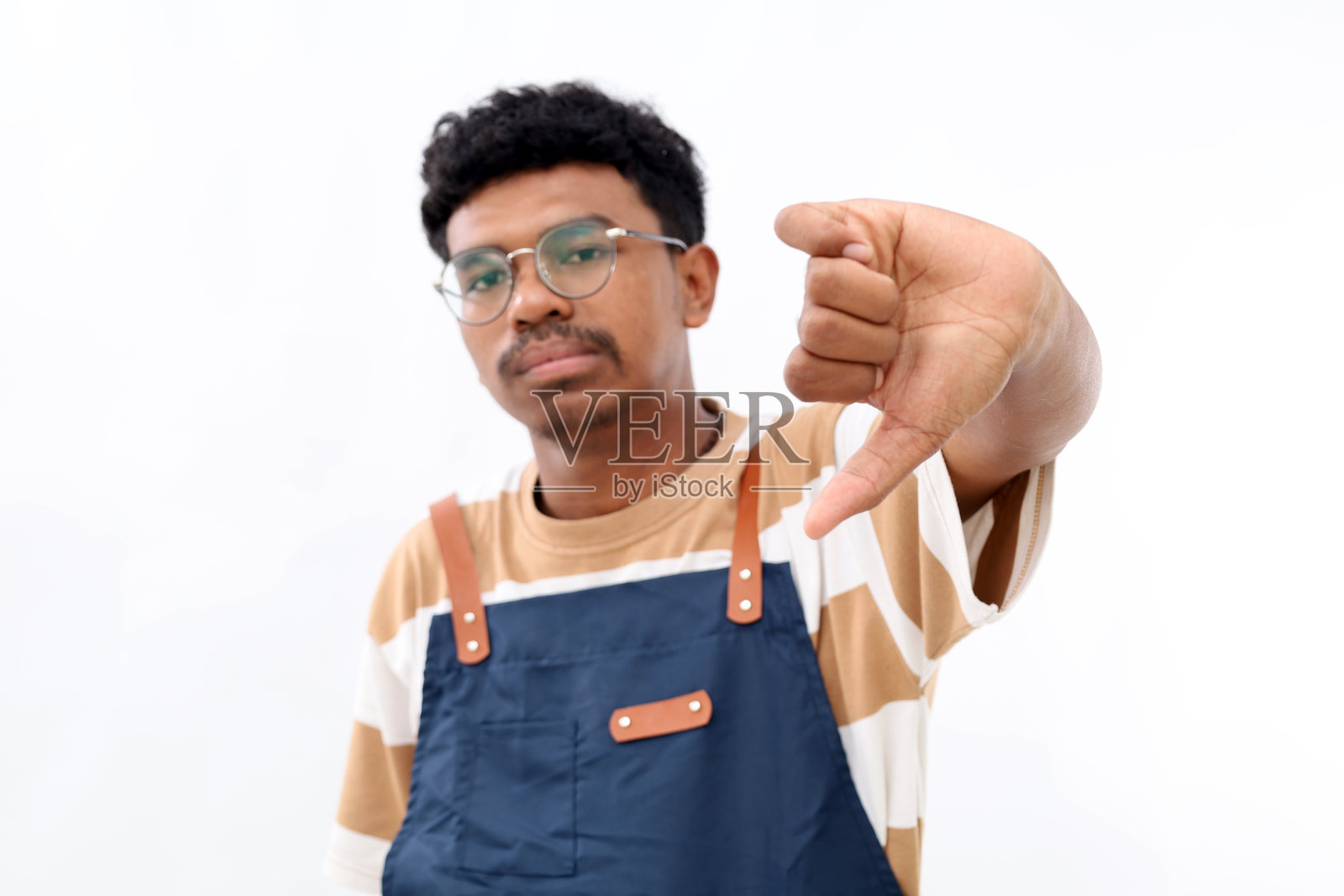 不满的亚洲男子穿着蓝色围裙,做出不赞同的手势,拇指向下,面露不悦,穿着红色衬衫,背景为白色,采用选择性聚焦。照片摄影图片