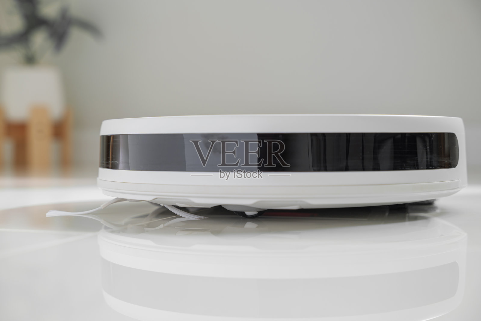 白色自动地面清洁机器人,配有侧刷,正在瓷砖地板上清洁,前方有黑色传感器条。机器人吸尘器高效清洁地面,展示智能家居技术。照片摄影图片