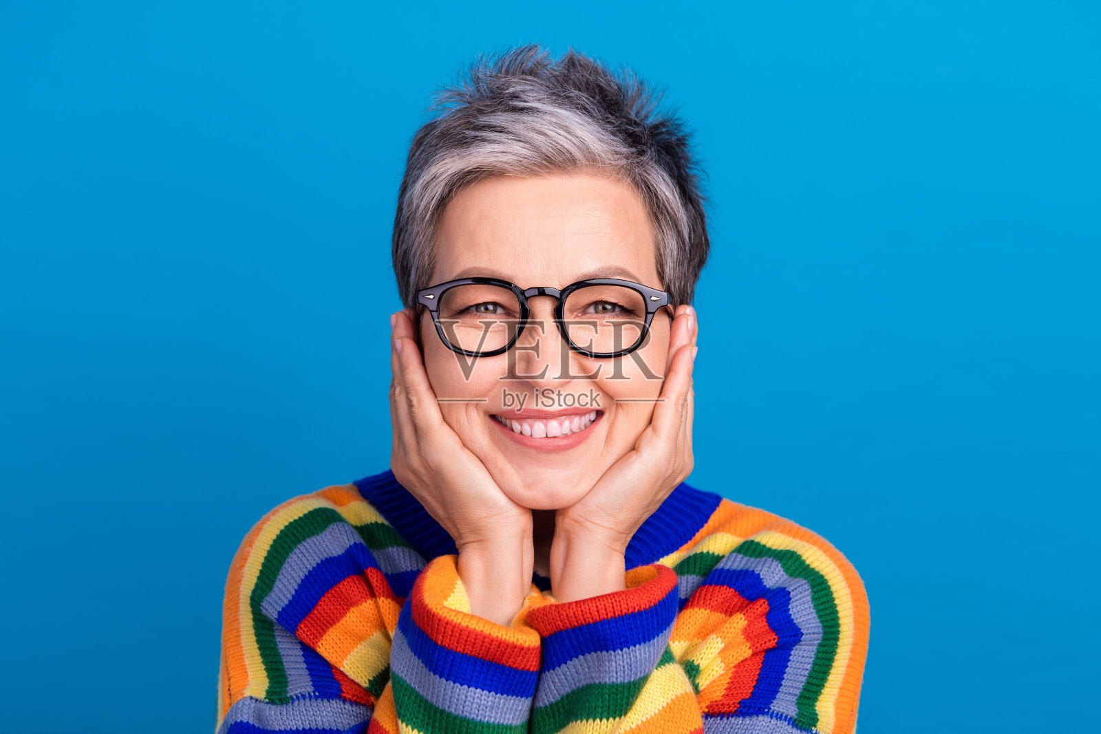 微笑的友好老年女士,双手托着脸颊,背景为蓝色。照片摄影图片