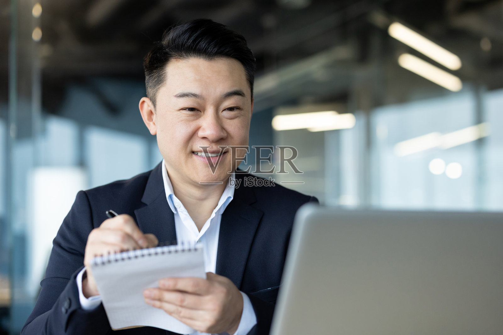 年轻亚洲男子穿着西装微笑,坐在办公室的工作场所,专注于笔记本电脑并在笔记本上做笔记的特写照片照片摄影图片