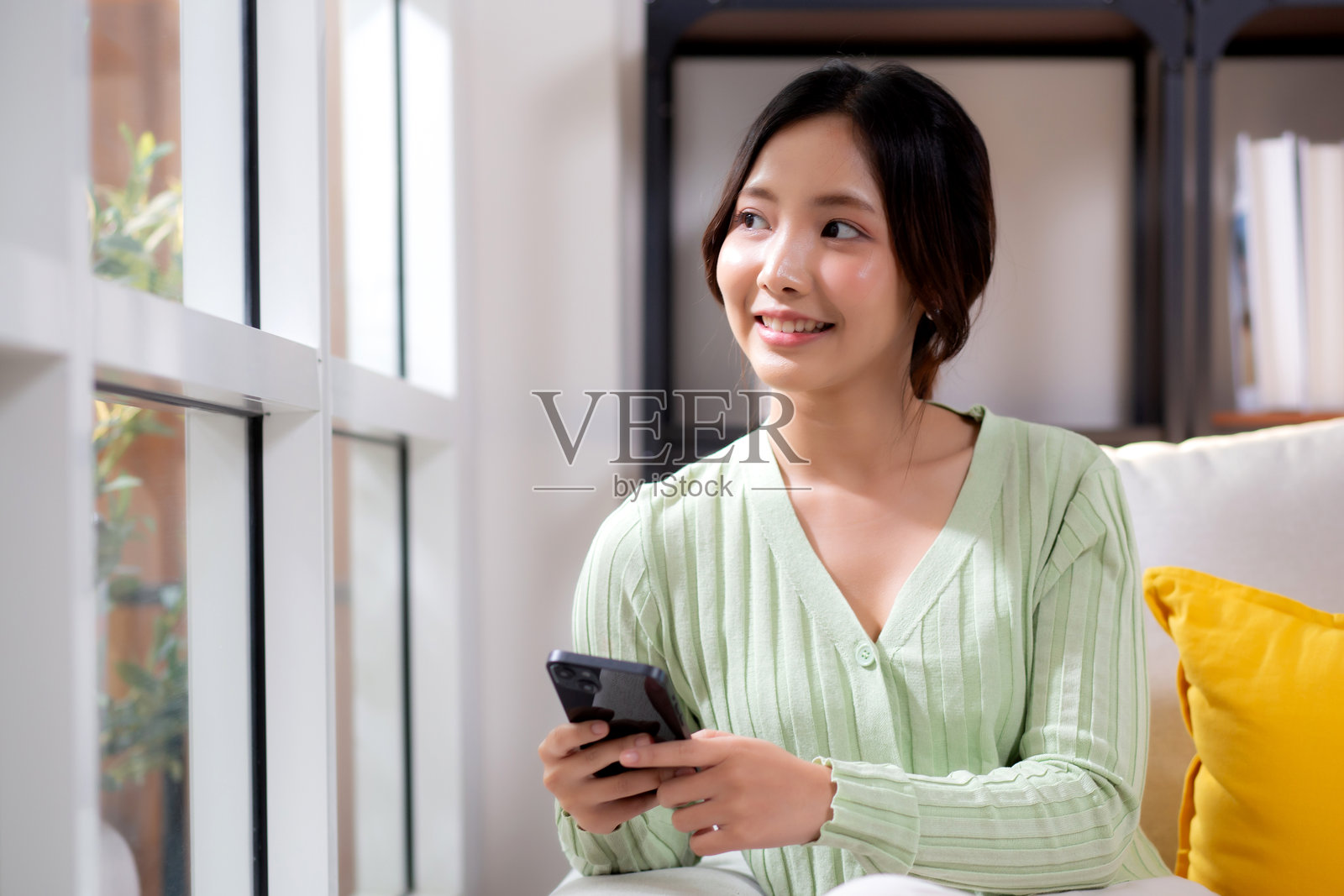 年轻的亚洲女商人坐在家里的沙发上,微笑着放松,查看智能手机,愉快的女性在家中沙发上观看智能手机,生活方式与沟通。照片摄影图片