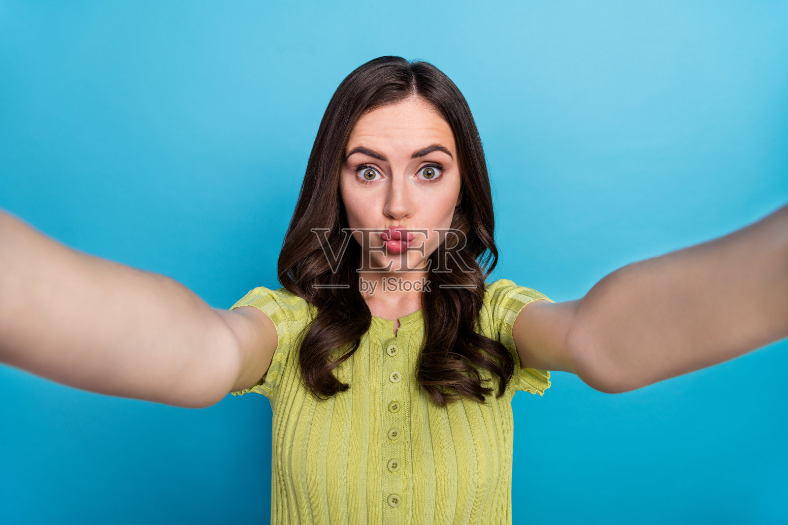 可爱的千禧一代棕发卷发自拍,送飞吻,穿着绿色上衣,背景为蓝色。照片摄影图片