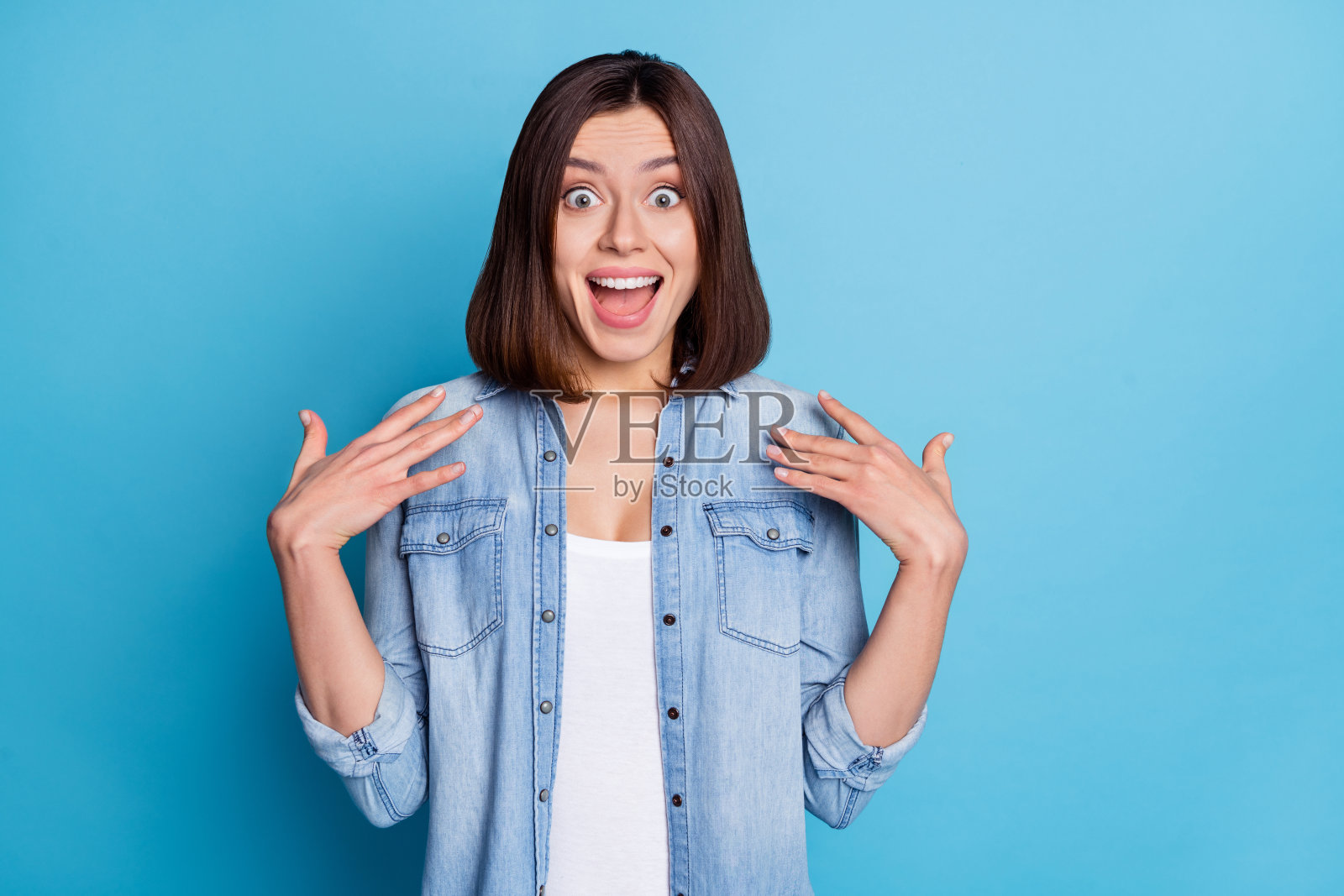 兴奋的甜美女子穿着牛仔衬衫,张开双臂,嘴巴大张,背景为蓝色。照片摄影图片