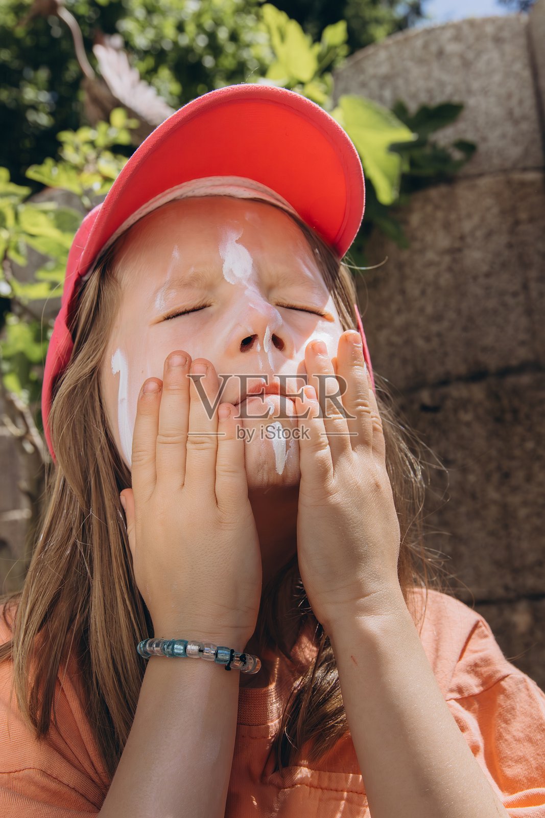 小女孩戴着粉色帽子,在阳光明媚的夏日为脸部涂抹防晒霜。护肤、阳光防护和儿童健康概念照片摄影图片